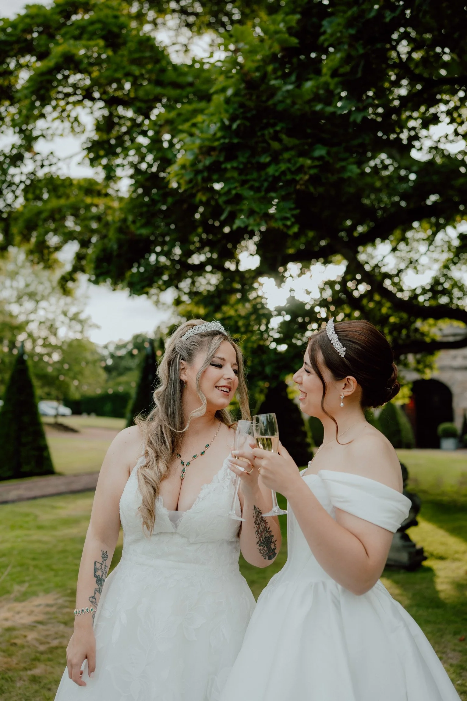 Same-sex newlyweds clinking glasses in the gardens at Prestonfield House, Edinburgh, during golden hour, captured in a romantic editorial wedding photography style.