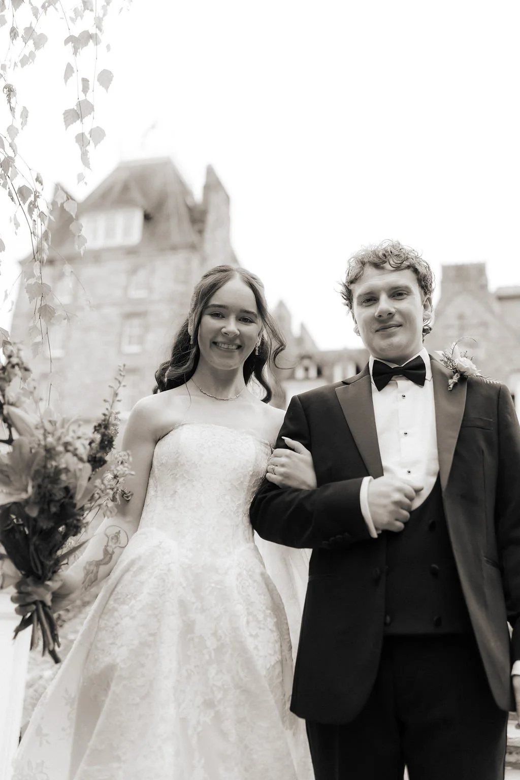 Black-and-white portrait of the smiling bride and groom standing arm in arm with Atholl Palace Hotel behind them.
