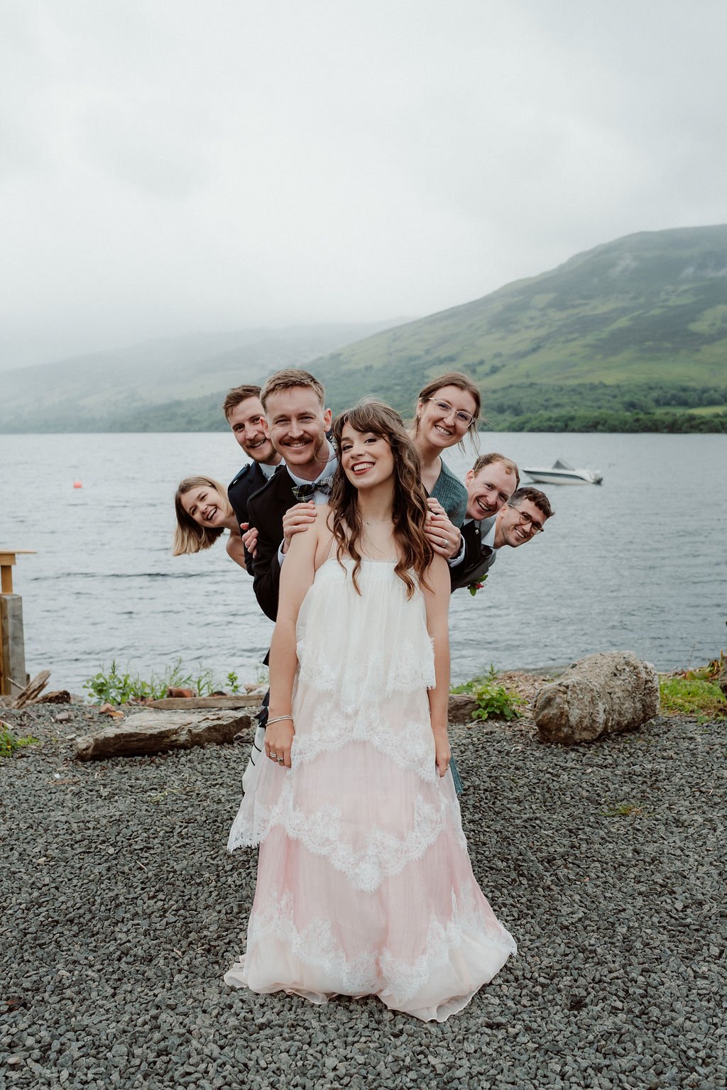 Bride smiling with her friends beside a Scottish loch, with misty Highland mountains in the background photographed by Patrice Little Photography.