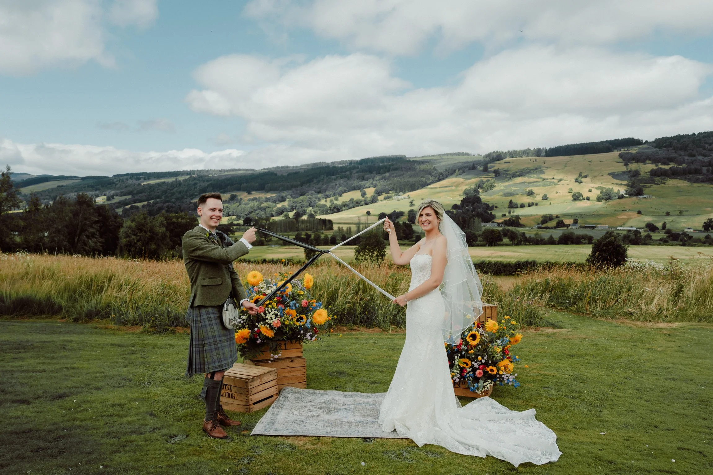Bride and groom celebrating their outdoor wedding ceremony at Ballintaggart Farm in Perthshire with colourful summer flowers, rolling Scottish hills and panoramic countryside views. Documentary wedding photography by Patrice Little Photography.