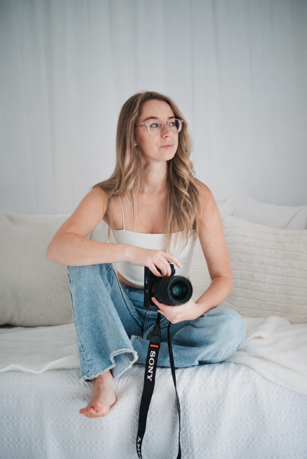 Woman with glasses sitting on bed with a camera, wearing a white tank top and jeans, in a minimalistic bedroom.