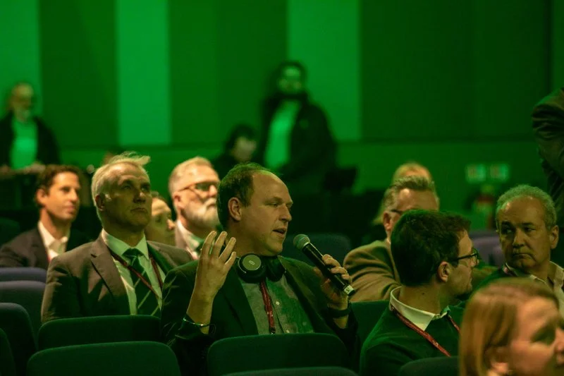 Man speaking into a microphone at a conference with audience members sitting in green-lit room.