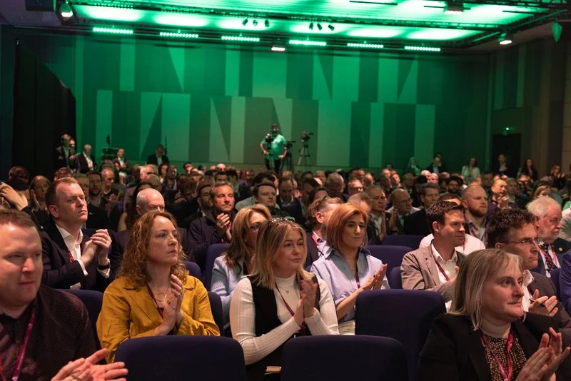 Audience seated in a conference room, paying attention to a presentation; green lighting and stage in the background.