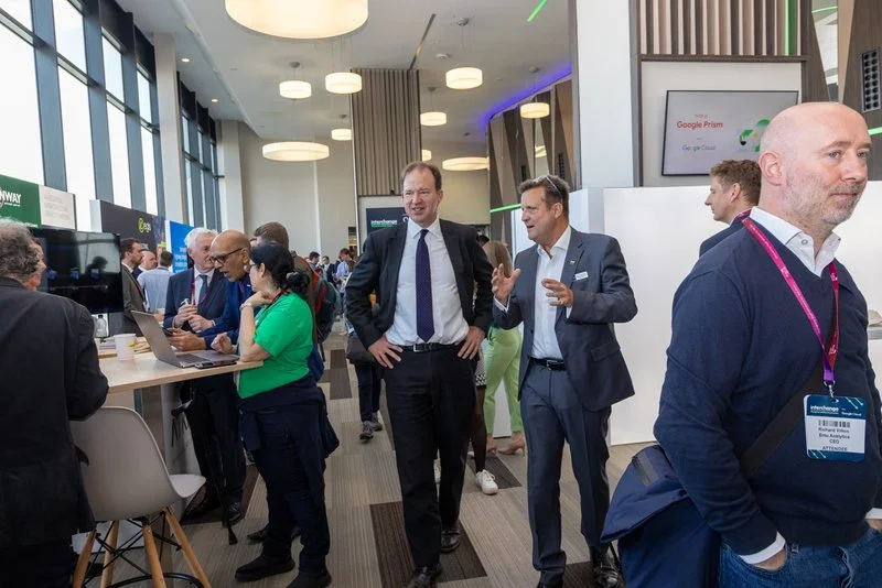 Business professionals at a conference or trade show, engaging in conversations and networking in a modern, well-lit venue with large windows and digital displays in the background.