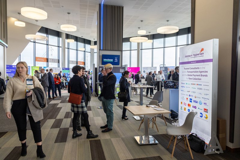 People attending a conference in a modern, well-lit hall with display booths and informational banners.