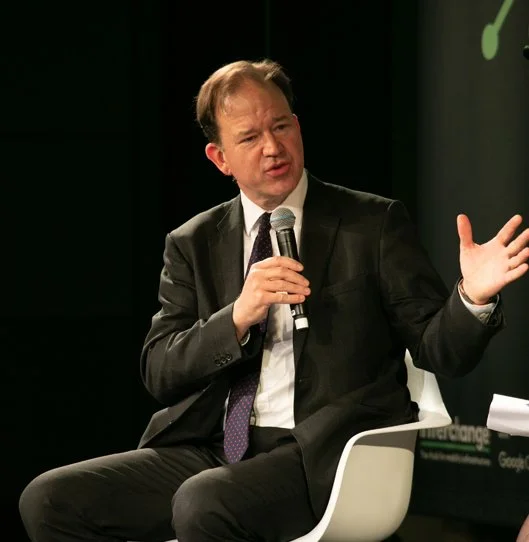 A man in a black suit, white shirt, and purple tie sitting on a white chair, holding a microphone, and gesturing with his other hand during a panel discussion or interview.