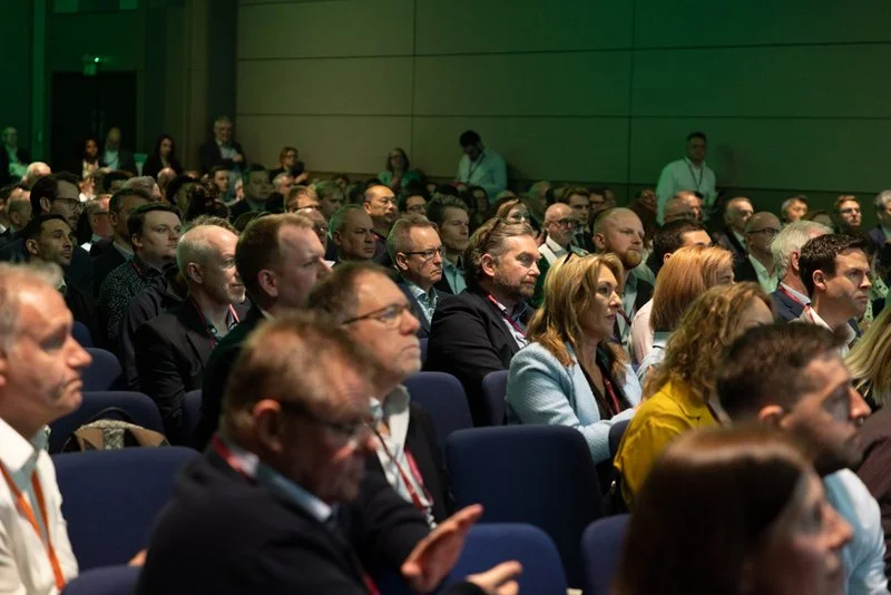 Audience of diverse professional individuals seated in a conference room, attentively listening to a presentation.