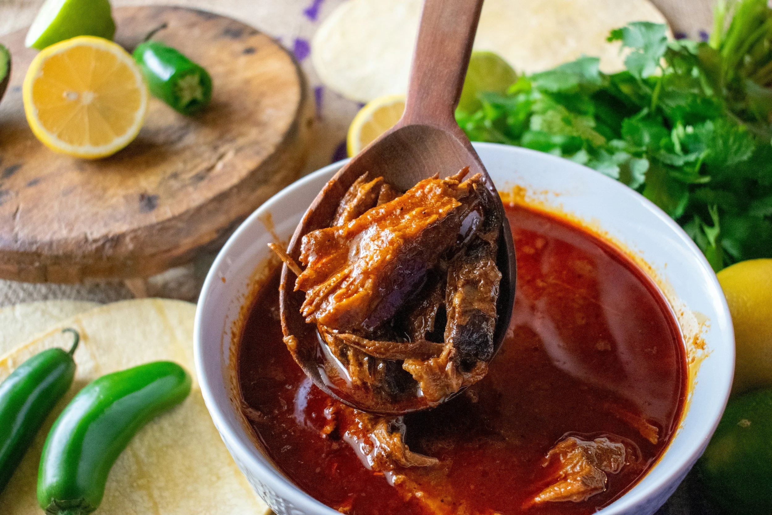 A bowl of a red soup with chunks of cooked beef, and herbs, lemon, and peppers in the background