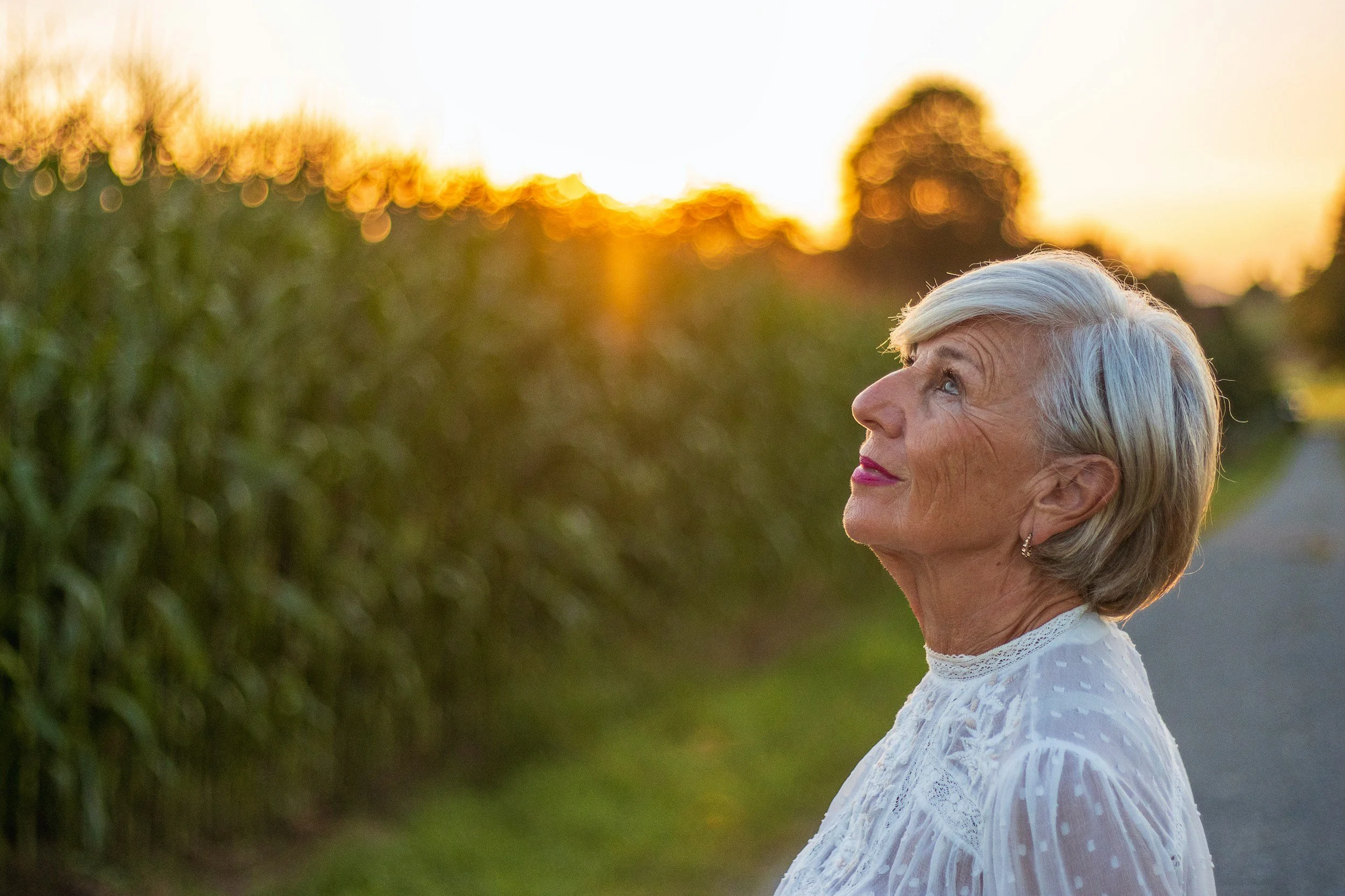 Woman with short gray hair and wearing a white lace top stands outdoors at sunset, looking up with a serene expression.