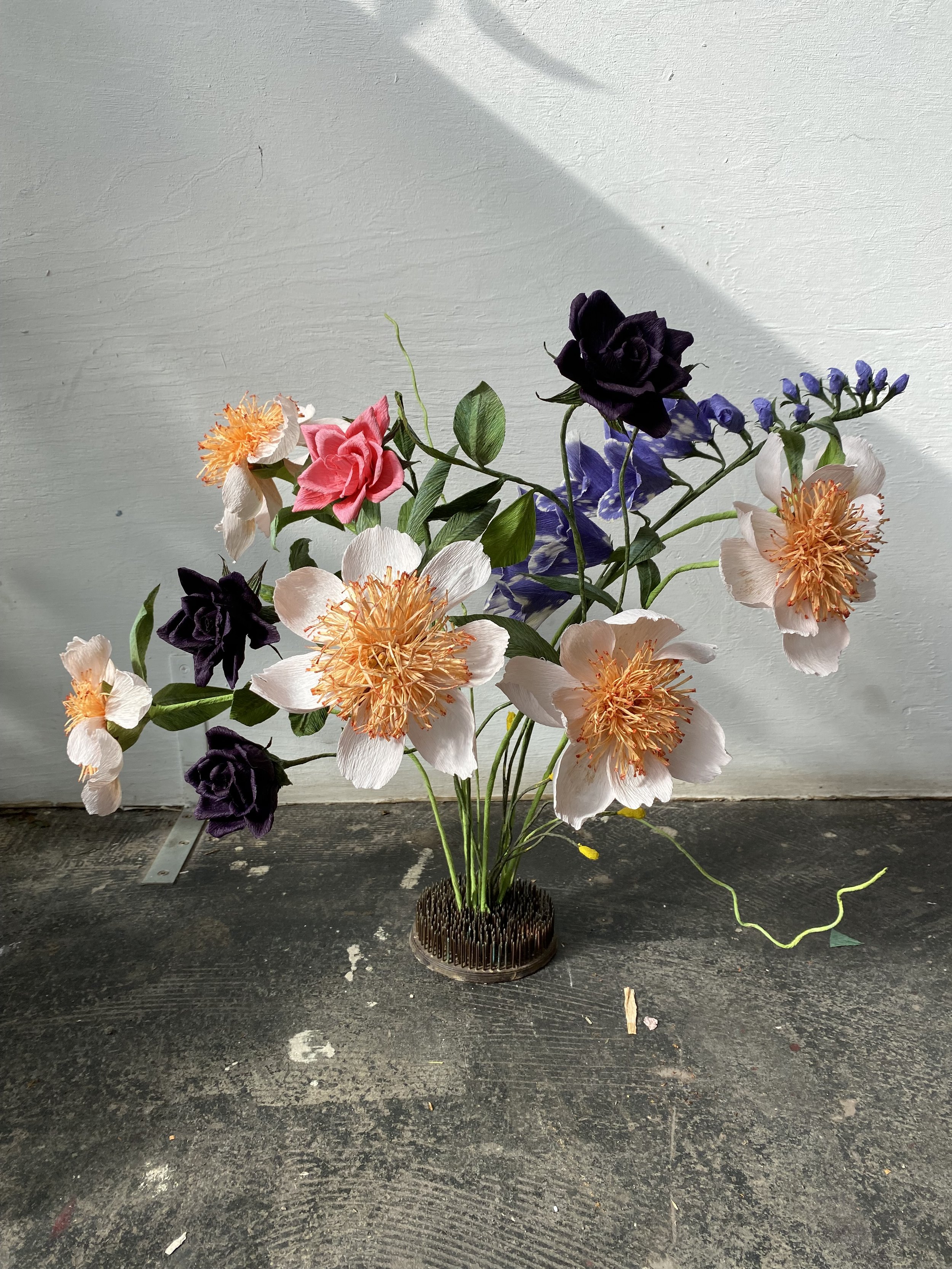 A bouquet of mixed artificial flowers by Emmeli Kimhi in a round black base, placed on a dark, worn wooden surface against a plain white wall with sunlight and shadow.