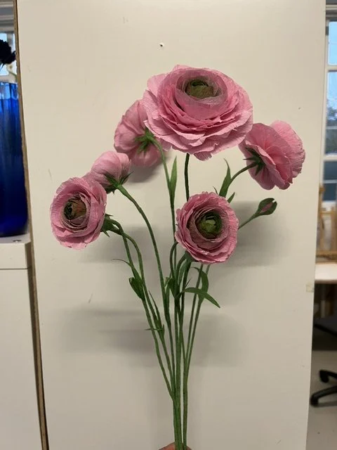 Paper pink flowers by Emmeli Kimhi with green stems and buds displayed against a plain background.