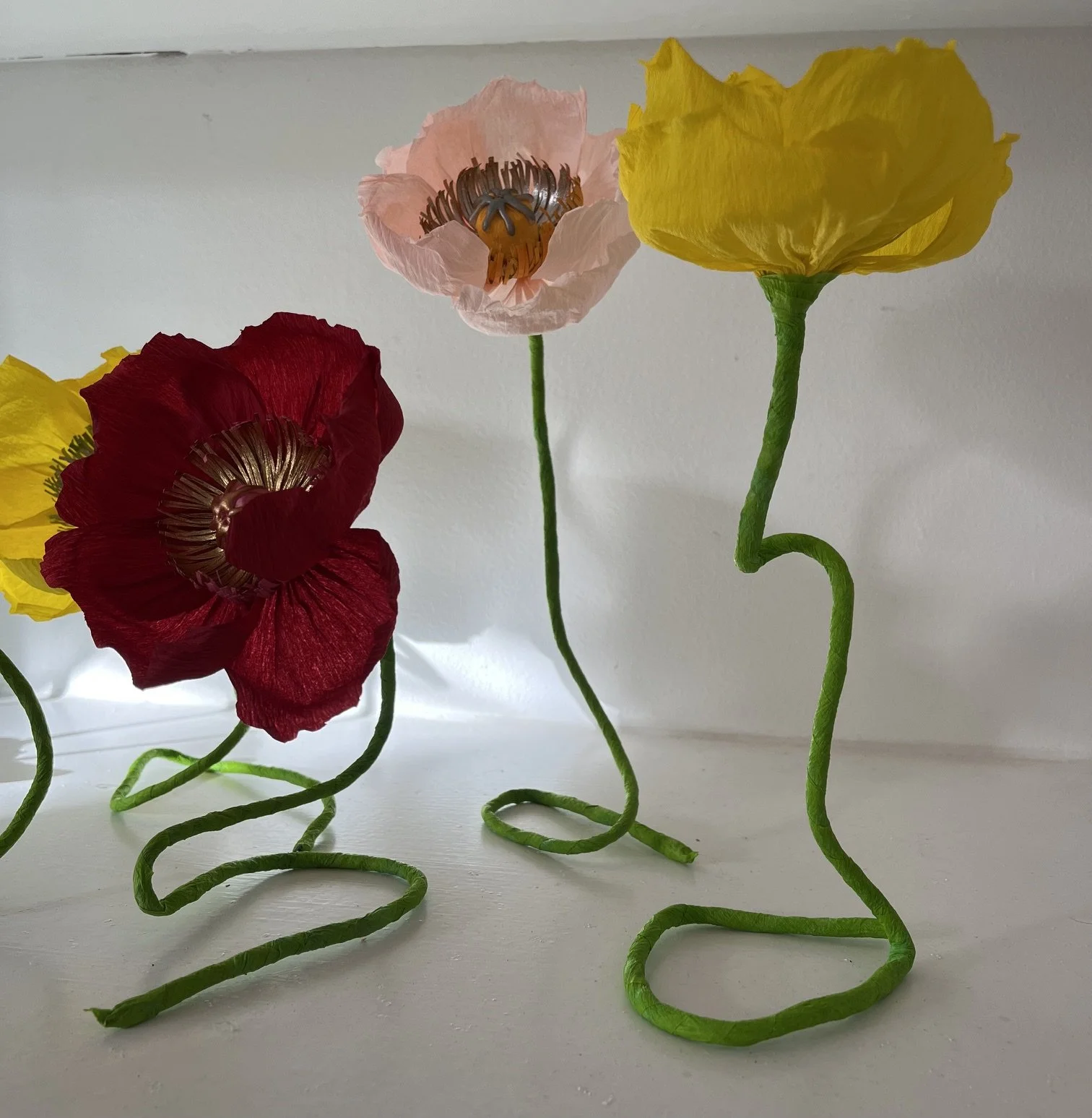 Colorful paper flowers with green twisted stems on a white surface by Emmeli Kimhi.