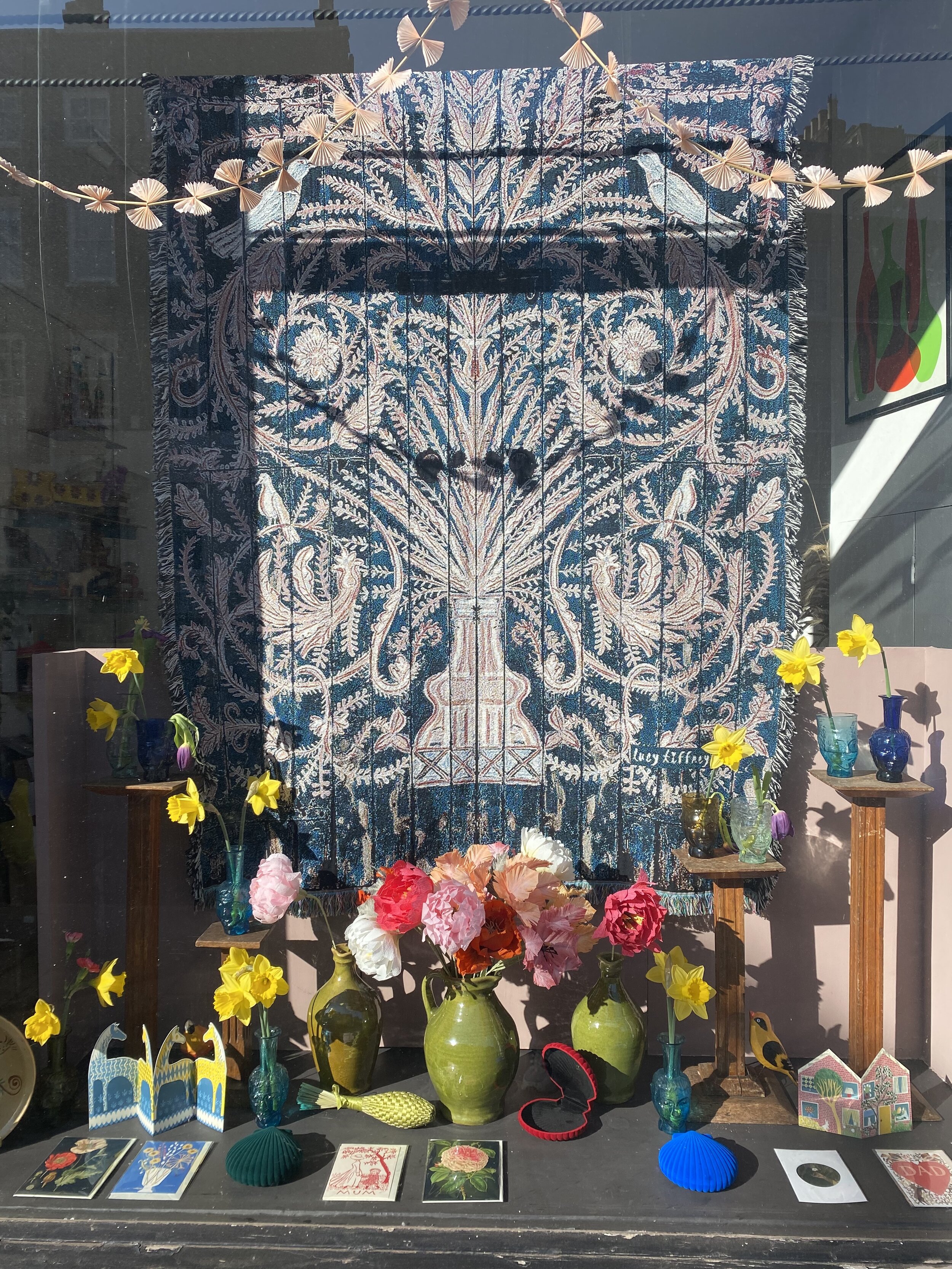 Display window featuring a floral art piece with a dark blue background and intricate designs, surrounded by vases of colorful flowers, paper fans, and various decorative objects. Paper flowers by Emmeli Kimhi