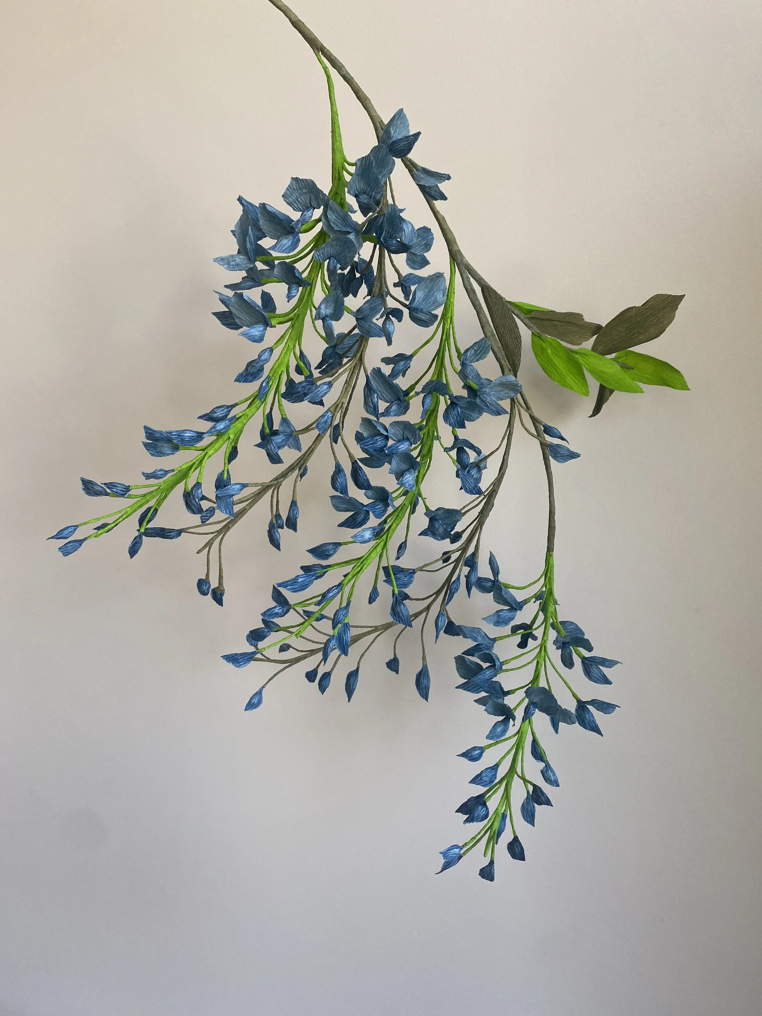 Artificial blue flowers by Emmeli Kimhi with green leaves and brown stems on a white background.