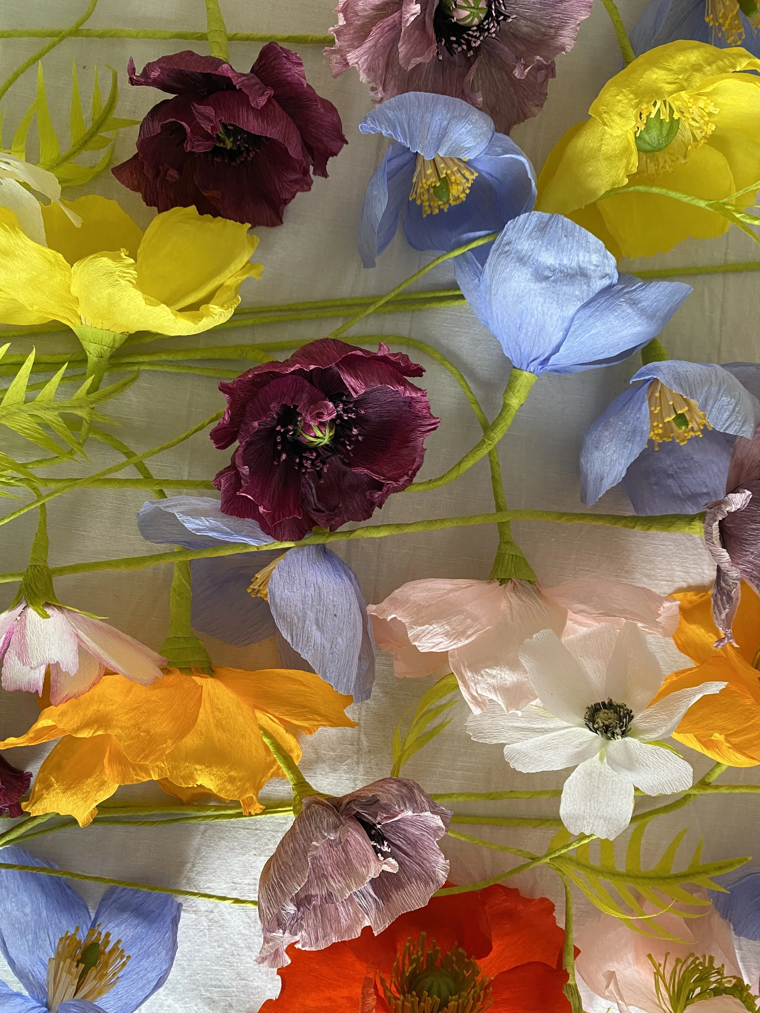 Colorful paper flowers by Emmeli Kimhi in various colors including yellow, purple, pink, white, orange, and blue, arranged with green paper leaves on a light background.
