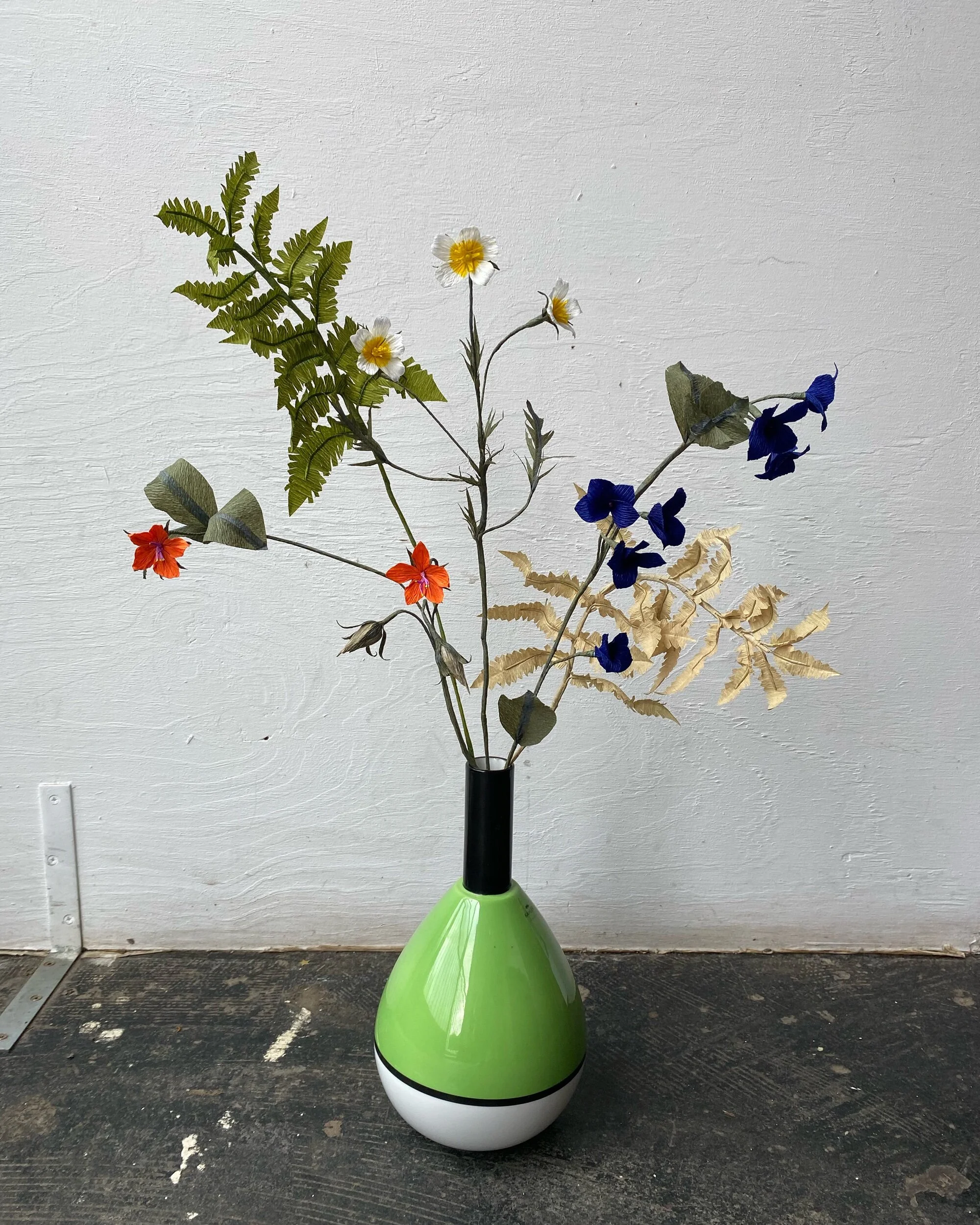 A decorative arrangement of faux flowers by Emmeli Kimhi and leaves in a black and green vase, placed on a dark, textured surface against a plain white wall.