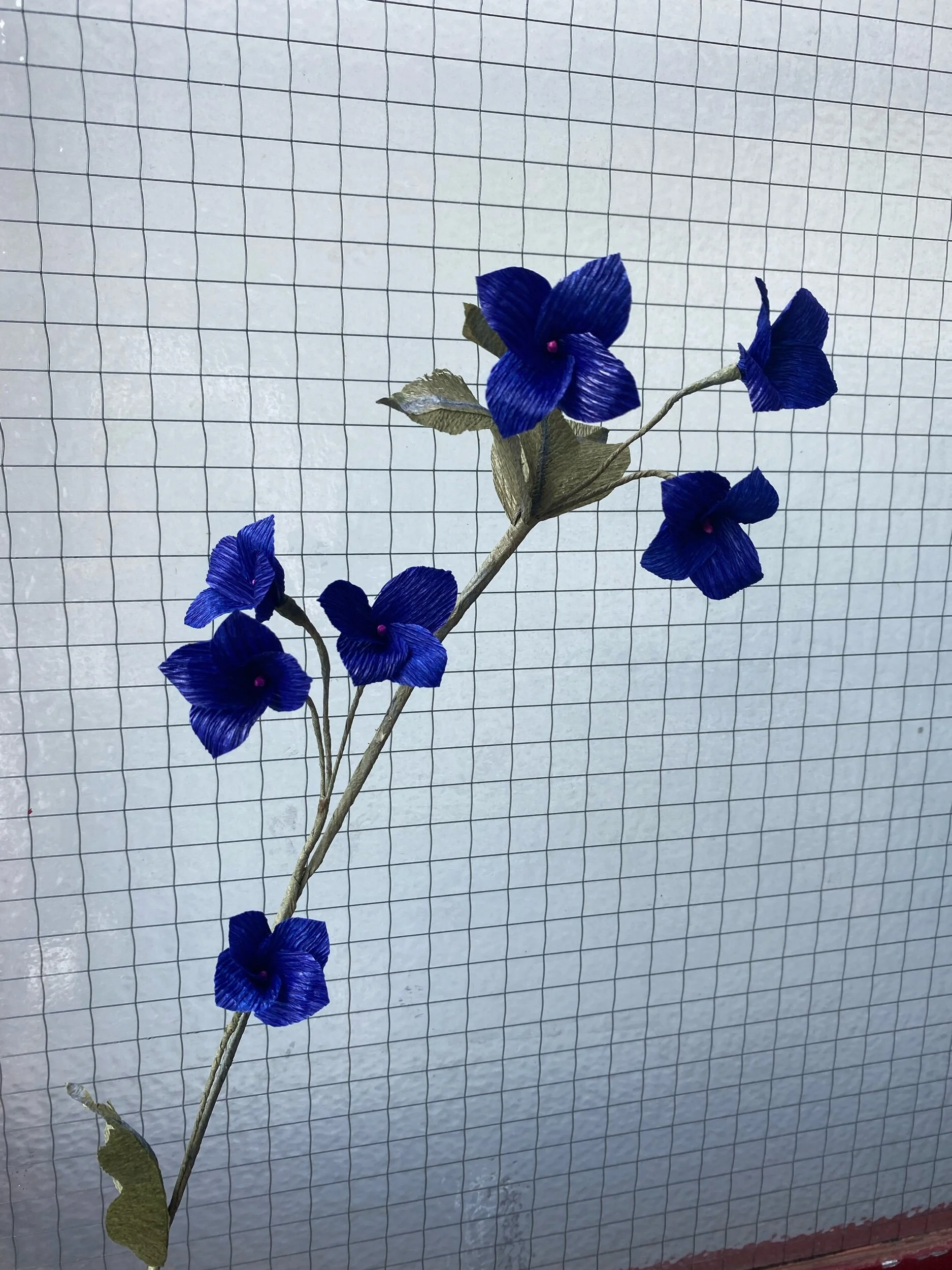 Artificial blue flowers with green leaves by Emmeli Kimhi on a branch against a wire grid background.