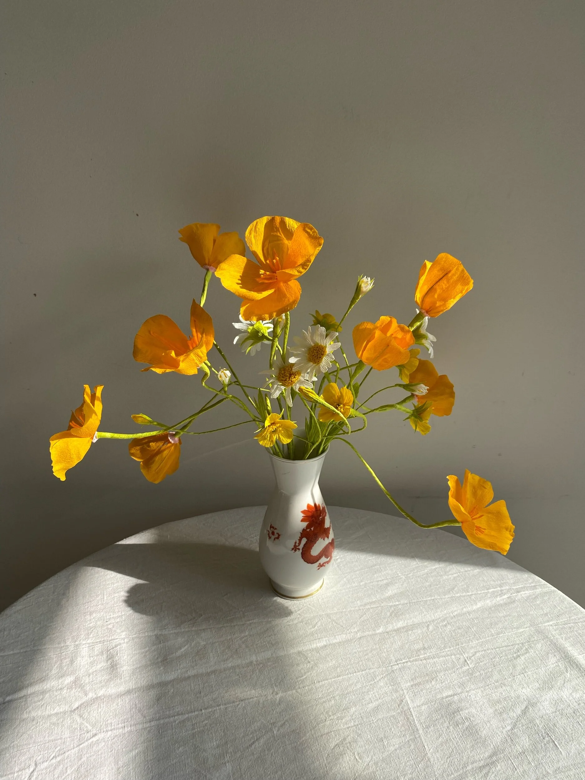 A white ceramic vase with red dragon design, holding a bouquet of yellow and white flowers by Emmeli Kimhi, placed on a white surface.