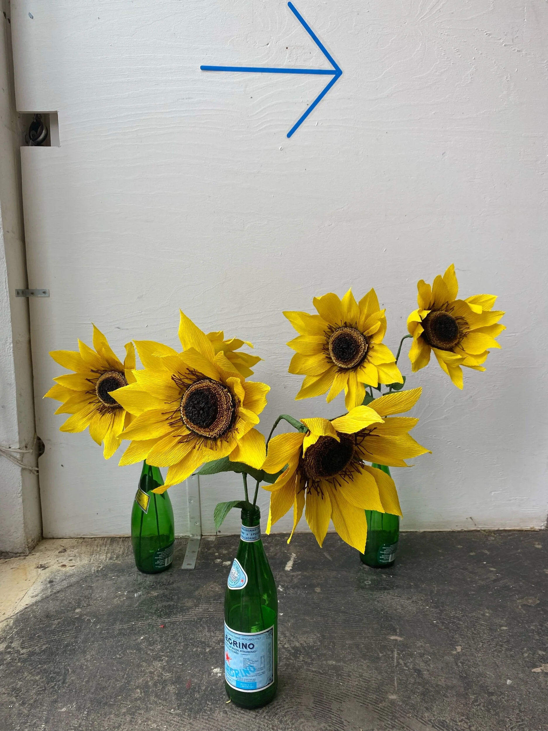 Three green bottles of sparkling mineral water with artificial sunflowers by Emmeli Kimhi attached to the necks, placed on a floor against a white textured wall with a blue arrow drawn on it.