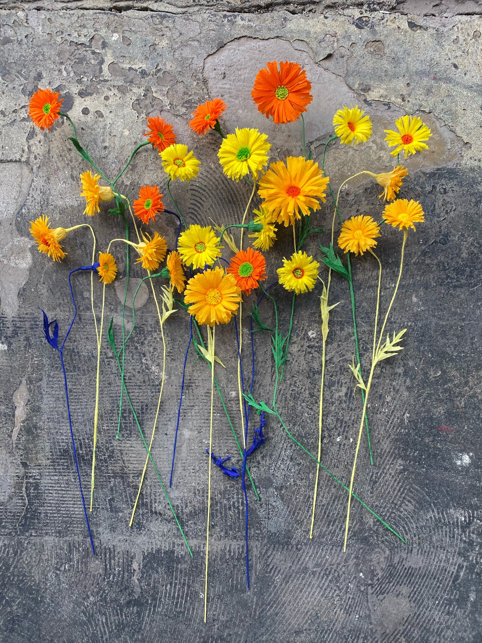 Colorful paper flowers with green, yellow, orange, and blue stems by Emmeli Kimhi arranged on a rough concrete ground.