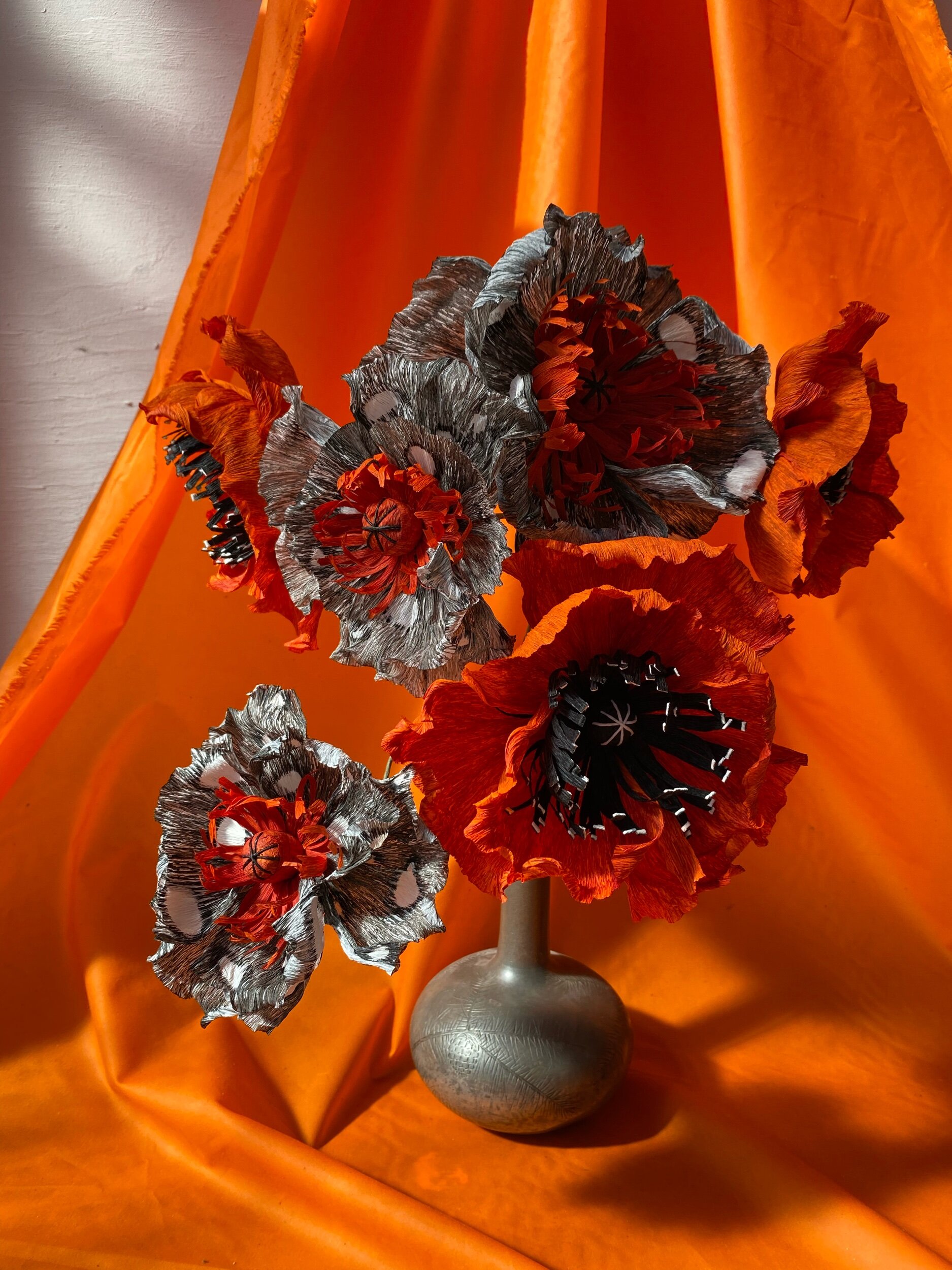 Artificial silver and red poppy flowers by Emmeli Kimhi in a silver vase with orange satin fabric background.