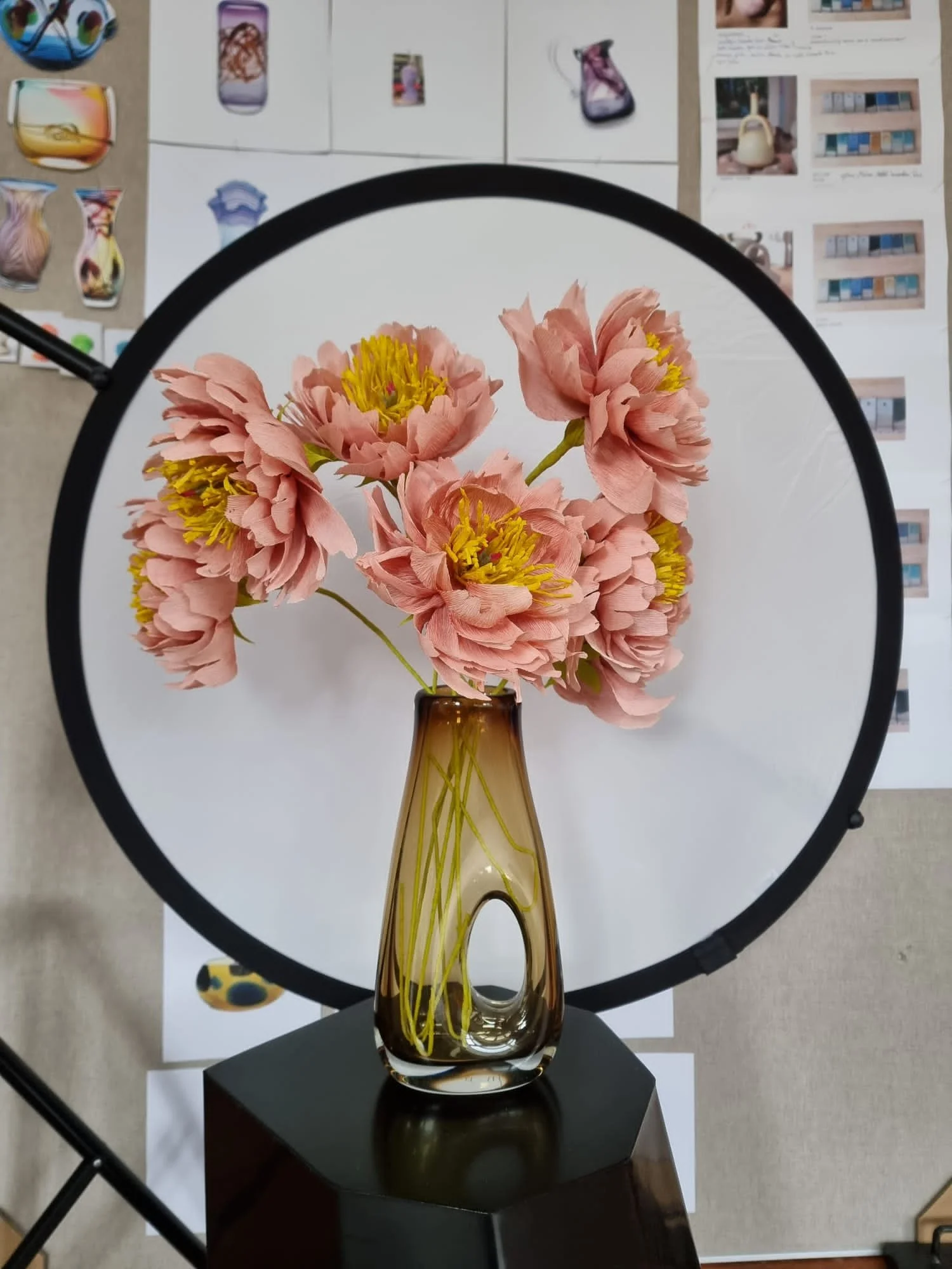 Close-up of a clear glass vase with pink artificial flowers by Emmeli Kimhi with yellow centers, reflected in a mirror ring, set on a black hexagonal table.