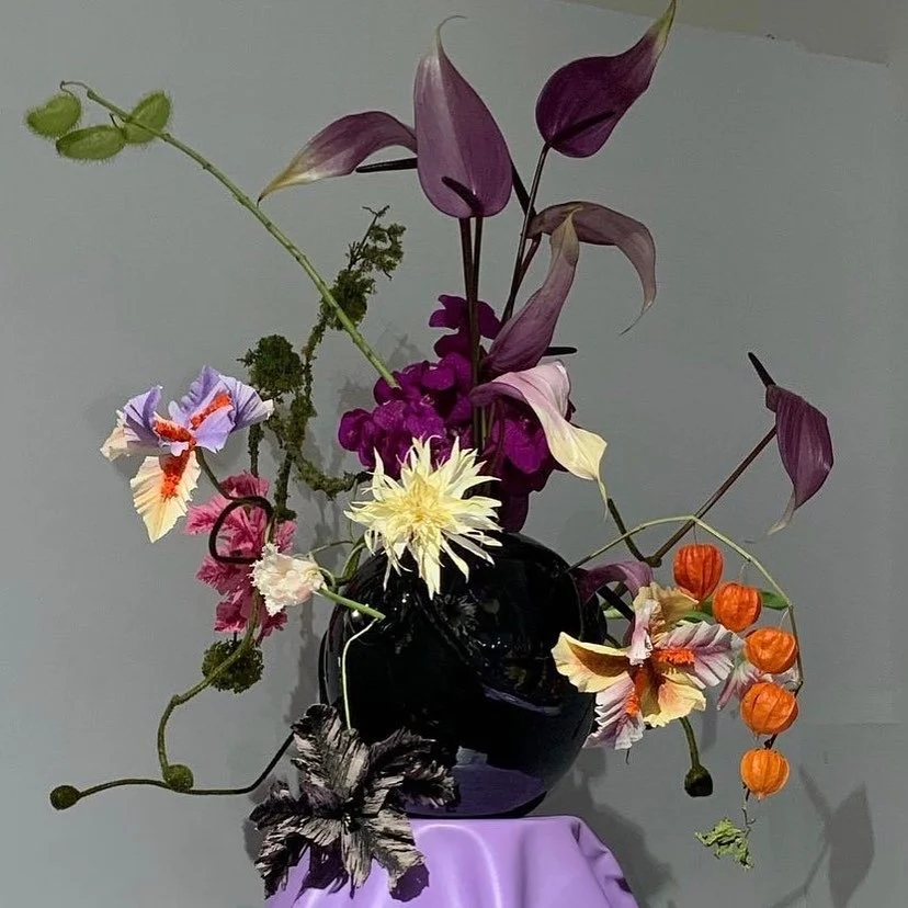 A floral arrangement in a black vase featuring purple, yellow, white, pink, and orange flowers with green leaves, placed on a lavender cloth against a gray background. Paper flowers by Emmeli Kimhi