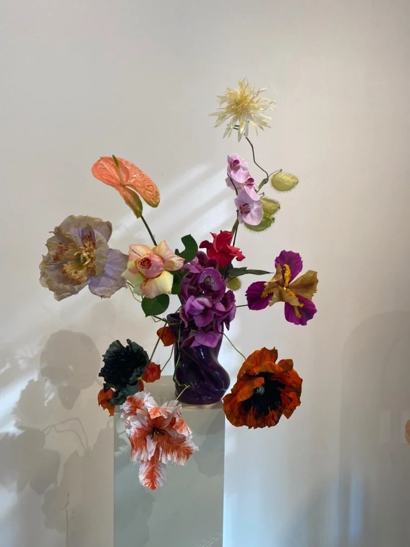 Colorful floral arrangement with various flowers in a purple vase on a white pedestal against a plain wall. Paper flowers by Emmeli Kimhi