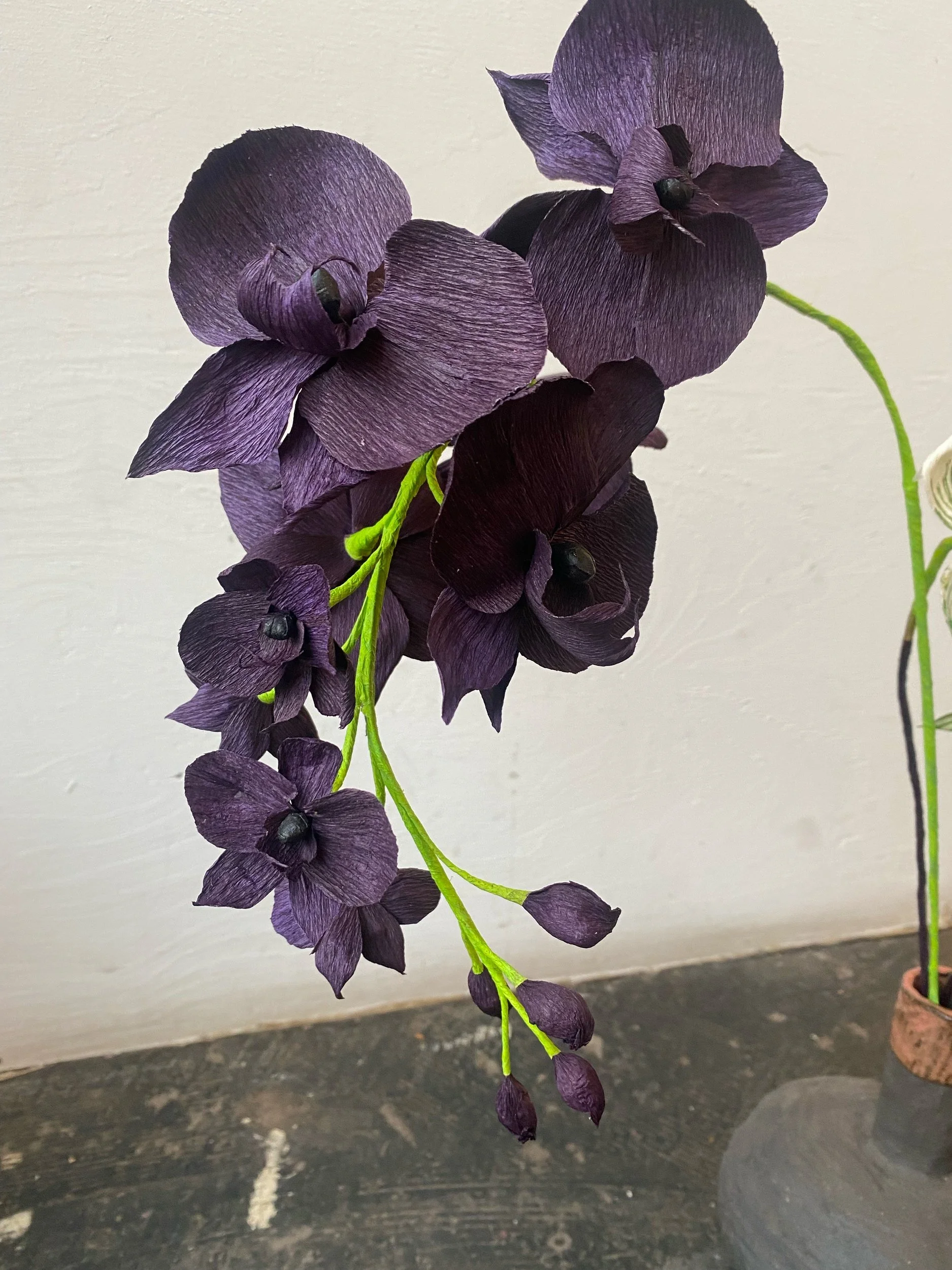 Purple  paper orchid flower  by Emmeli Kimhi with hanging petals and green stem against a plain light background.
