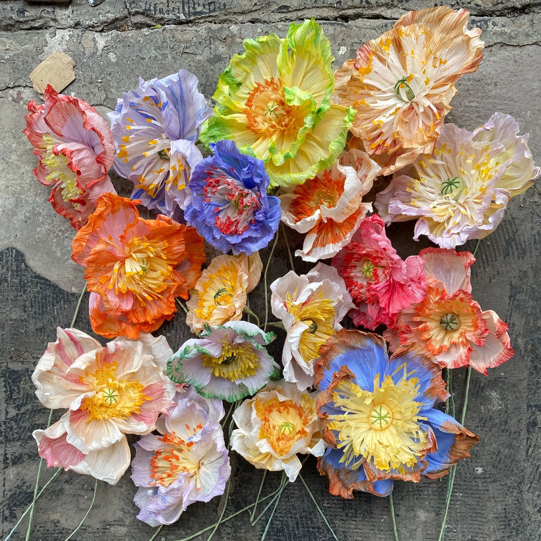 Colorful paper poppy flowers by Emmeli Kimhi arranged on a rough concrete surface.