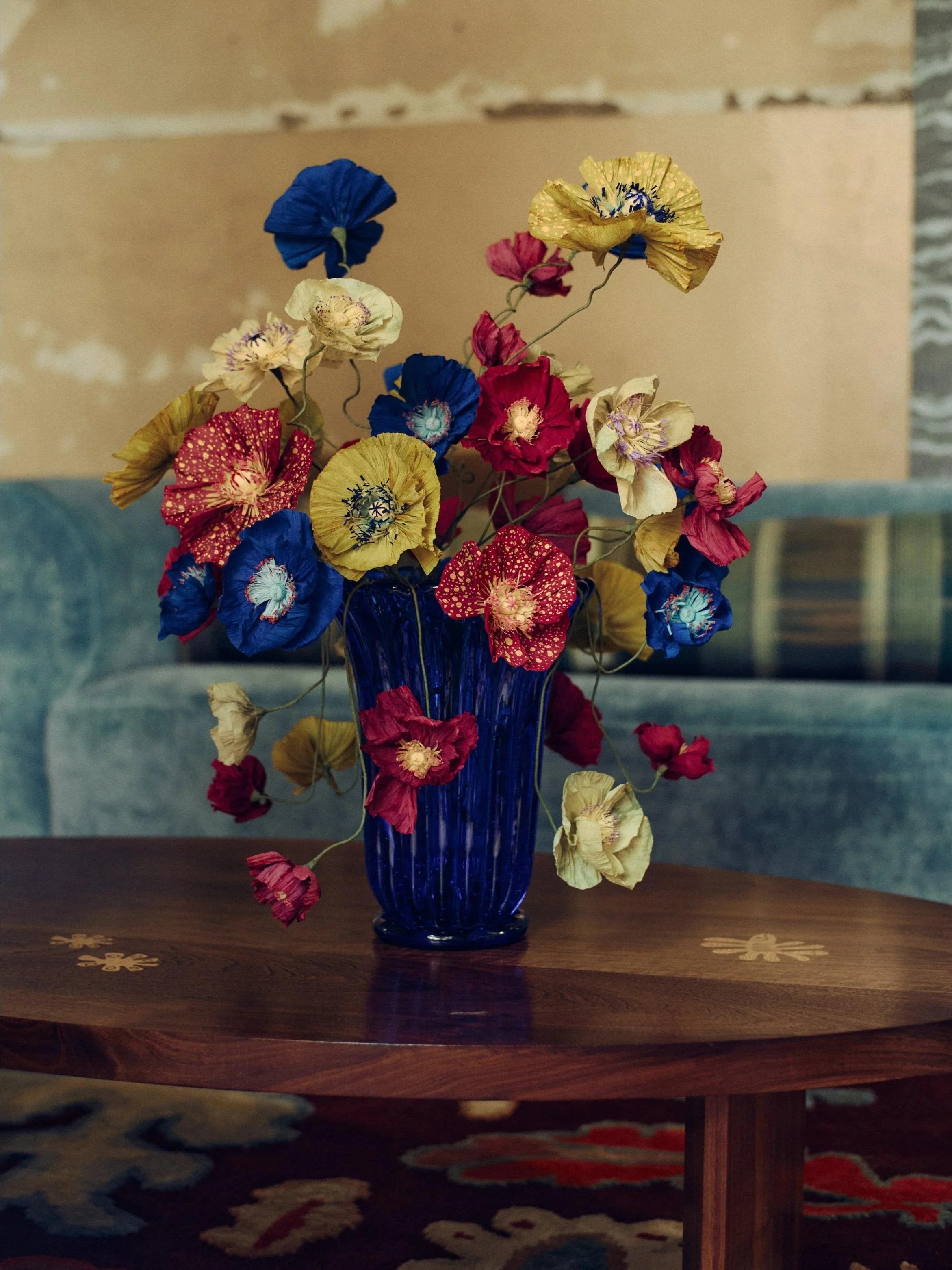 Handmade paper poppy bouquet created by Emmeli Kimhi for Sister by Studio Ashby, exhibited at Alcova Milan. Vibrant red, blue, and yellow paper flowers in a deep blue glass vase.
