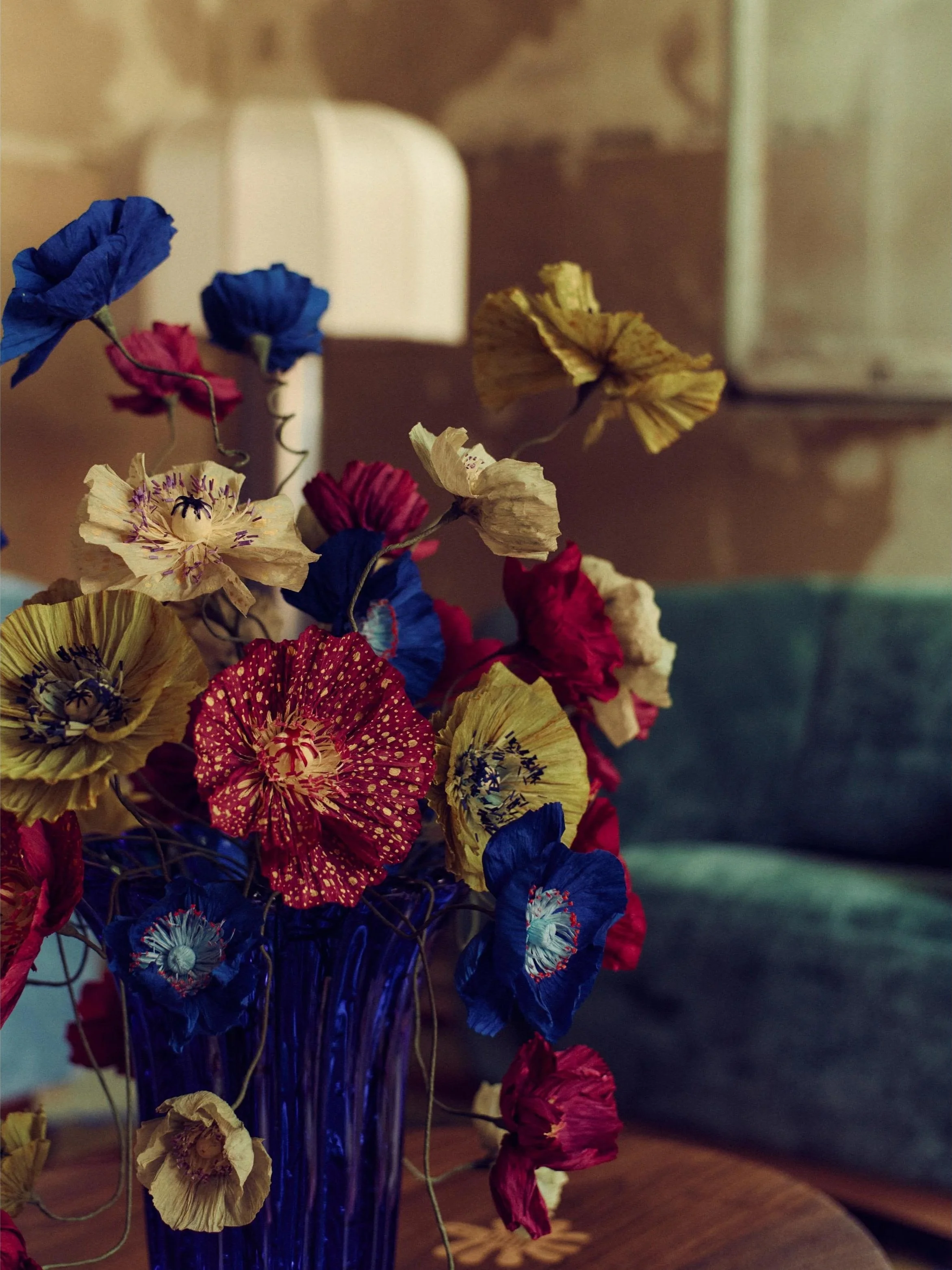 Handmade paper poppy bouquet by Emmeli Kimhi for Sister by Studio Ashby, exhibited at Alcova Milan. Vibrant red, blue, and yellow poppies arranged in a blue glass vase.