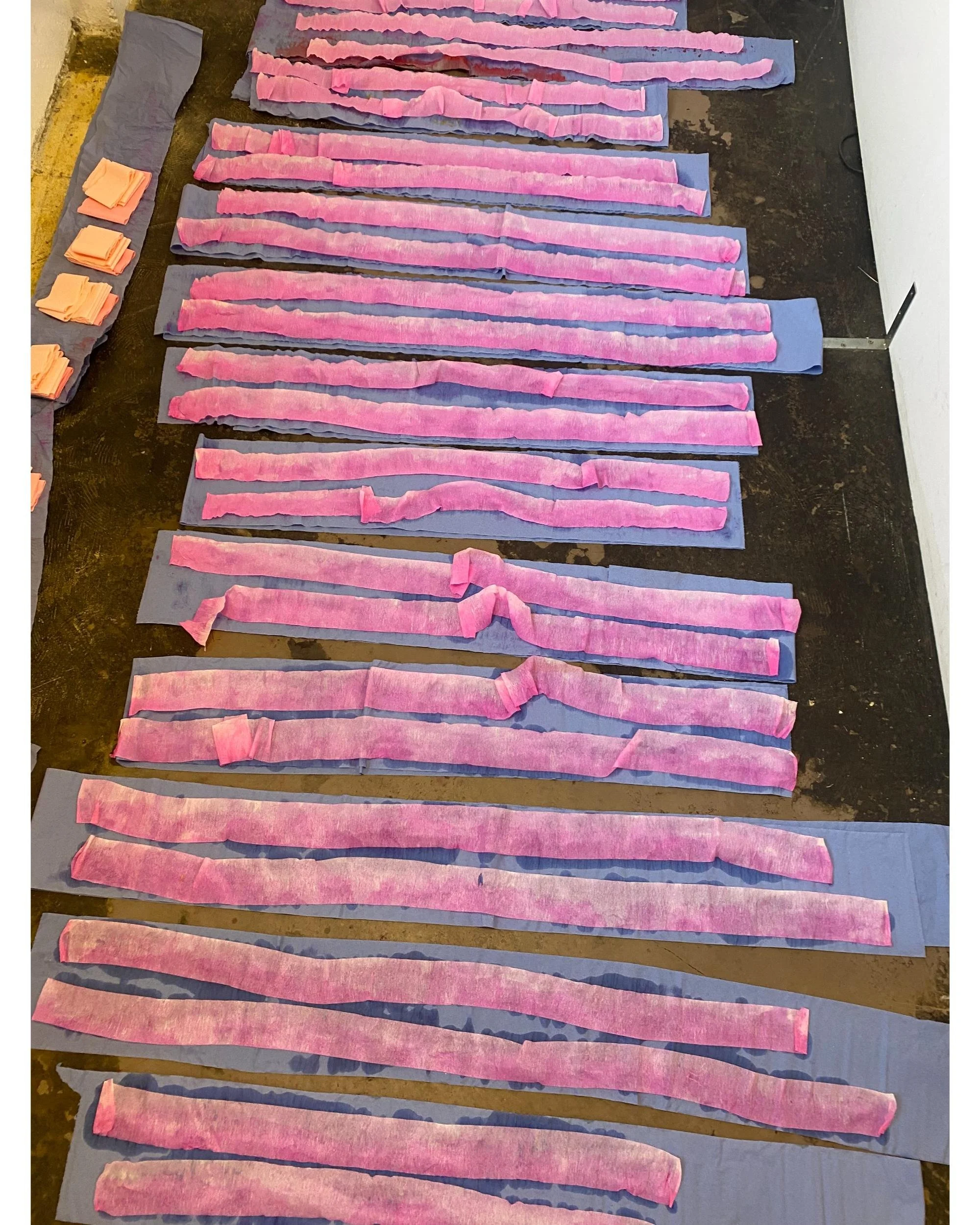 Multiple long strips of hand-dyed pink crepe paper in varying shades laid out on the studio floor, prepared for a large-scale paper sculpture by Emmeli Kimhi