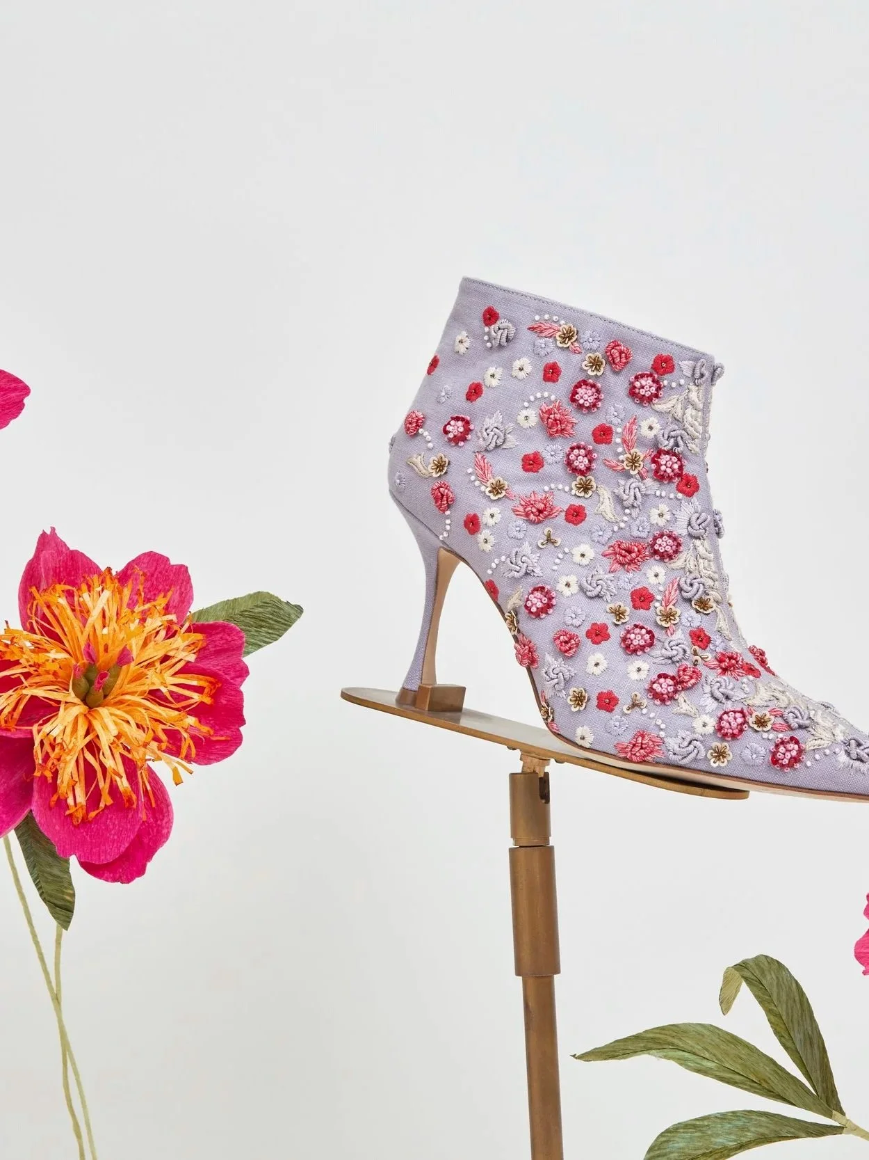 A high-heeled shoe with embroidery of red, pink, white, and lavender paper flowers by Emmeli Kimhi, positioned on a stand surrounded by pink and orange flowers against a white background.