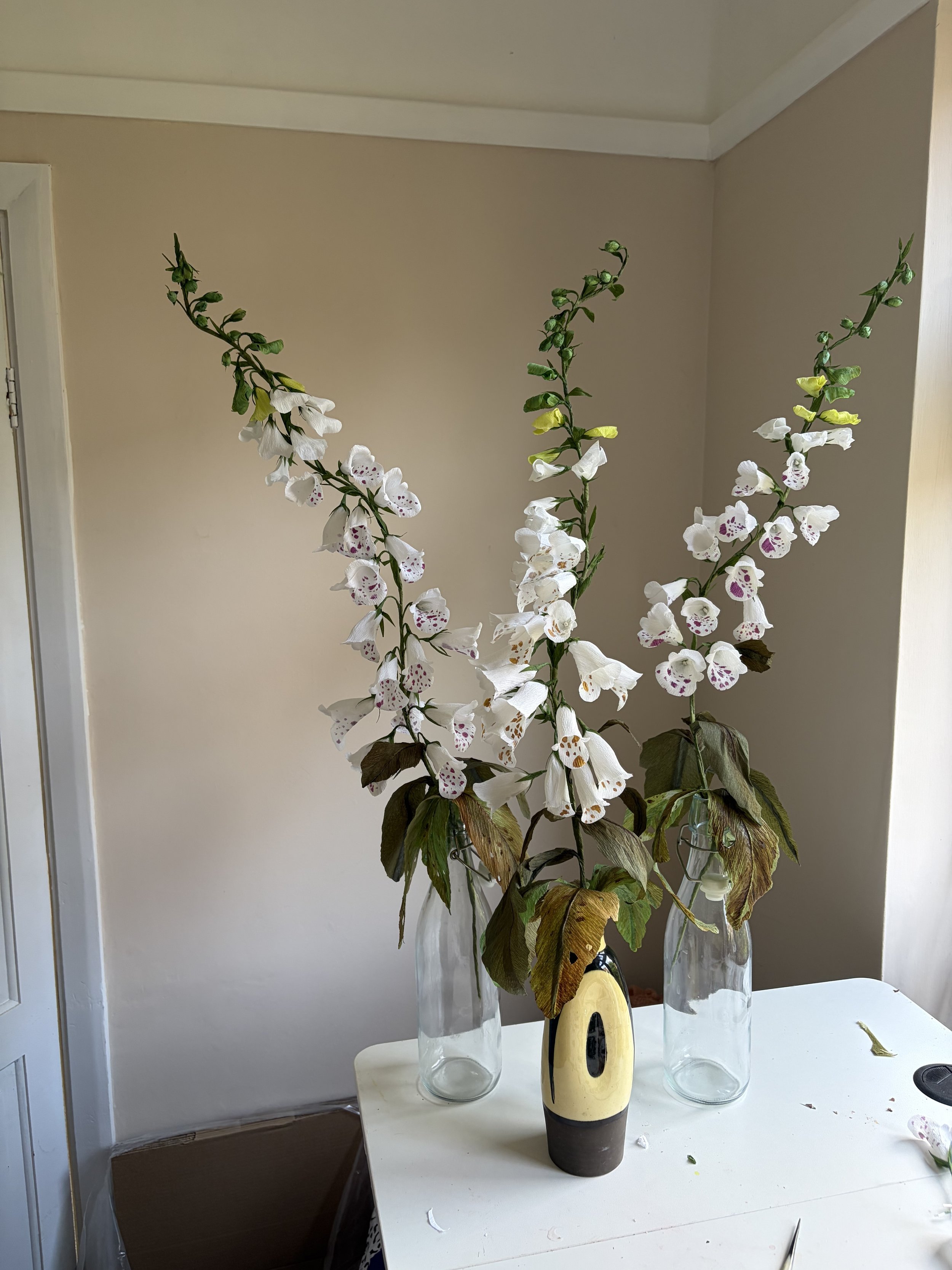 Three tall vases with crepe paper white foxgloves by Emmeli Kimhi and green leaves on a table, with a beige wall in the background.