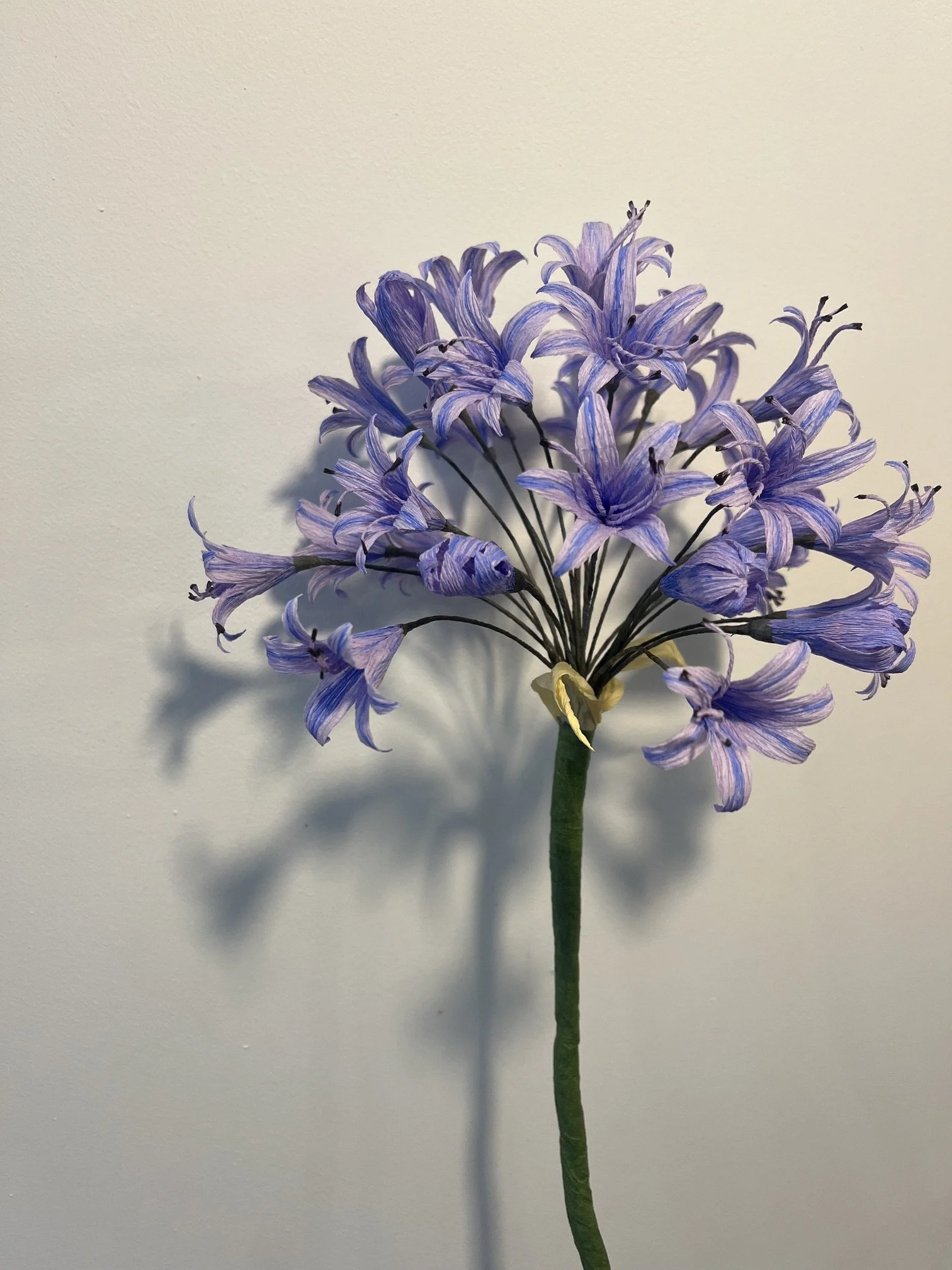 Fake purple flower bouquet by Emmeli Kimhi with a green stem against a plain light-colored wall.