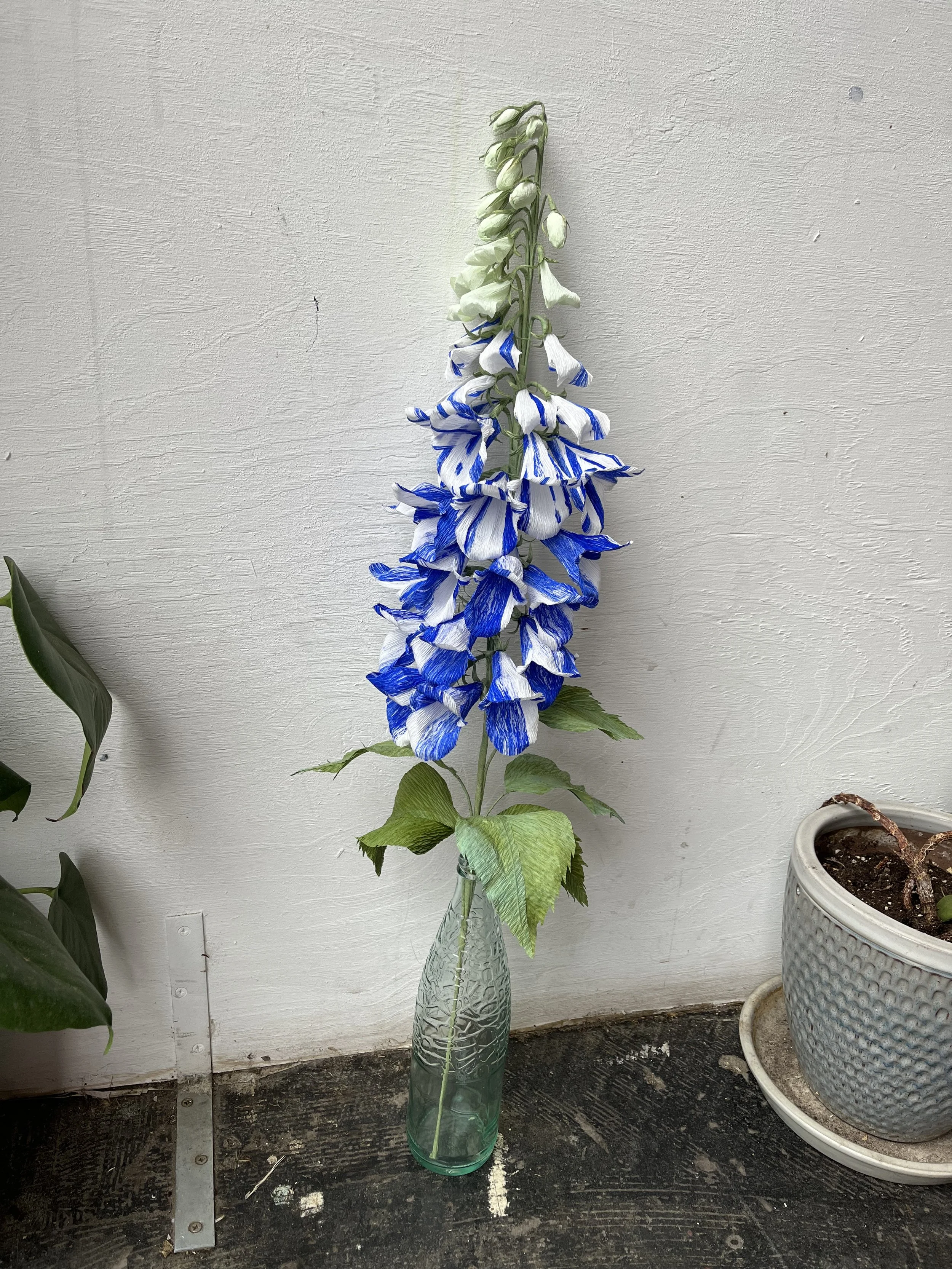 A tall green glass vase with a decorative pattern holds an artificial foxglove flower by Emmeli Kimhi with blue and white striped petals, positioned against a plain white wall, with a potted plant to the right and a dark wooden surface beneath.