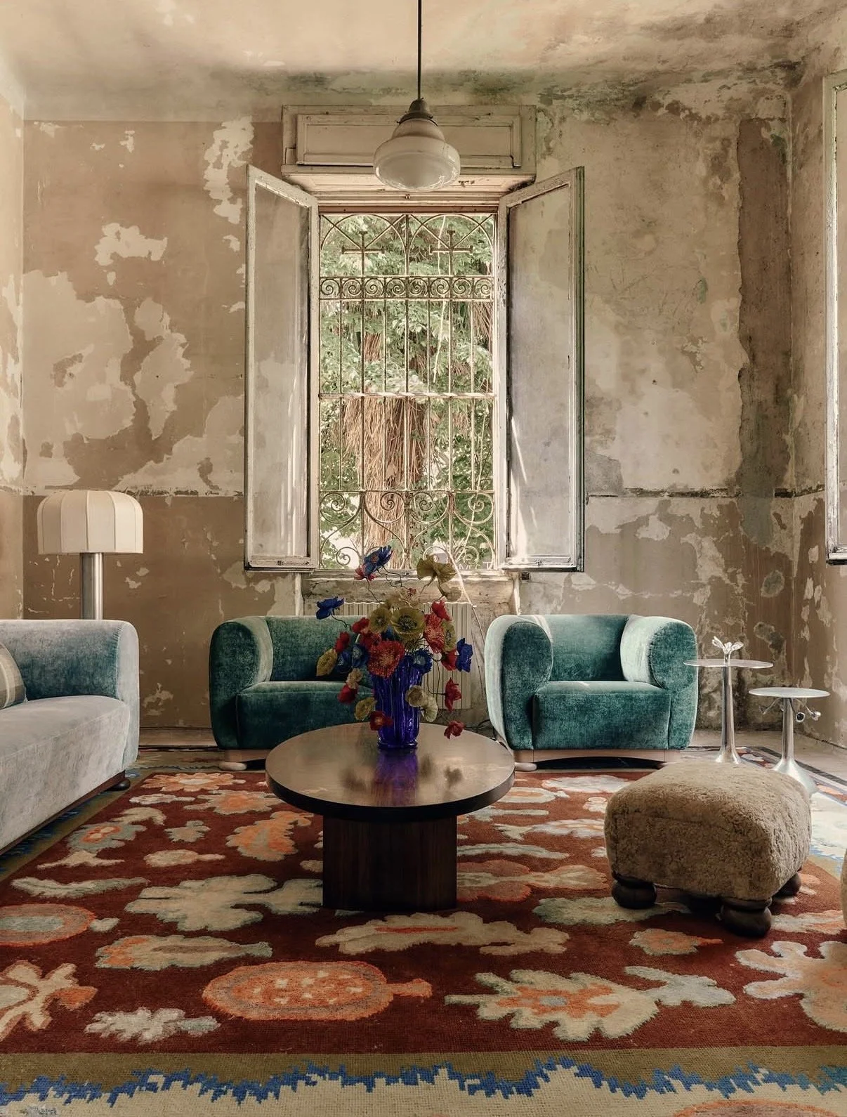 Emmeli Kimhi’s vibrant hand-painted paper poppy bouquet in a striking blue glass vase, centred on a round wooden coffee table in Studio Ashby’s installation at Alcova Milan. The colourful arrangement is styled in a room with distressed walls, teal ve