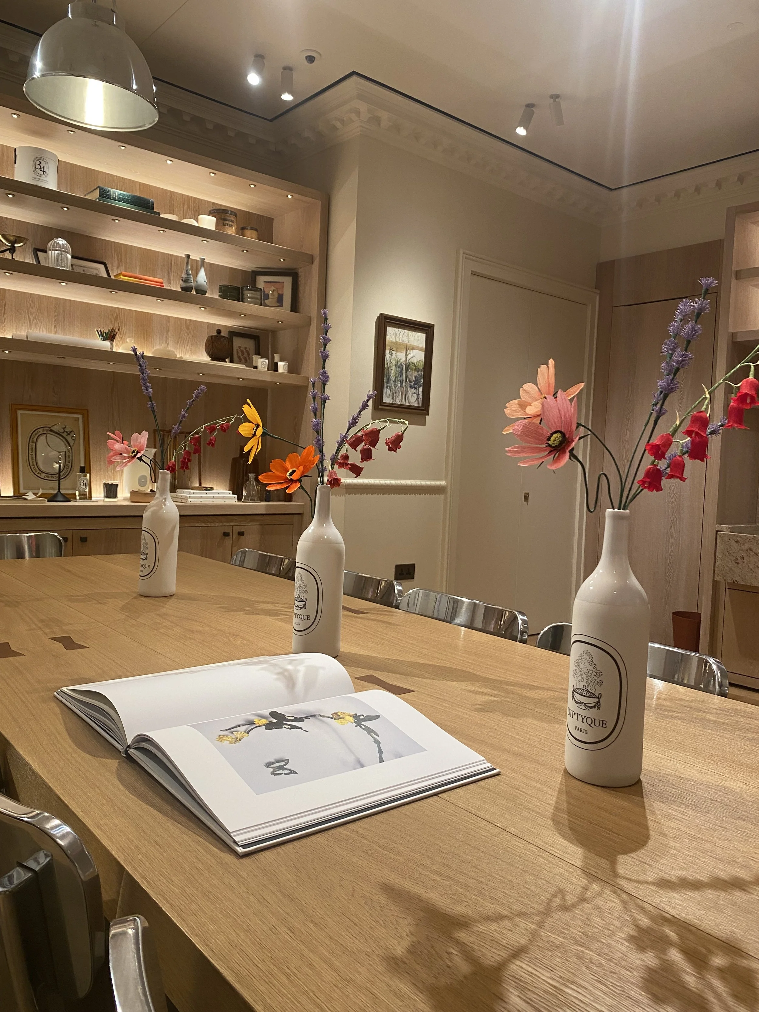 A wooden table with three white bottles containing colorful paper flowers by Emmeli Kimhi, an open book with illustrations, and some chairs in a well-lit room with wooden shelves and decorative items. At the Diptyque store on Bond Street.