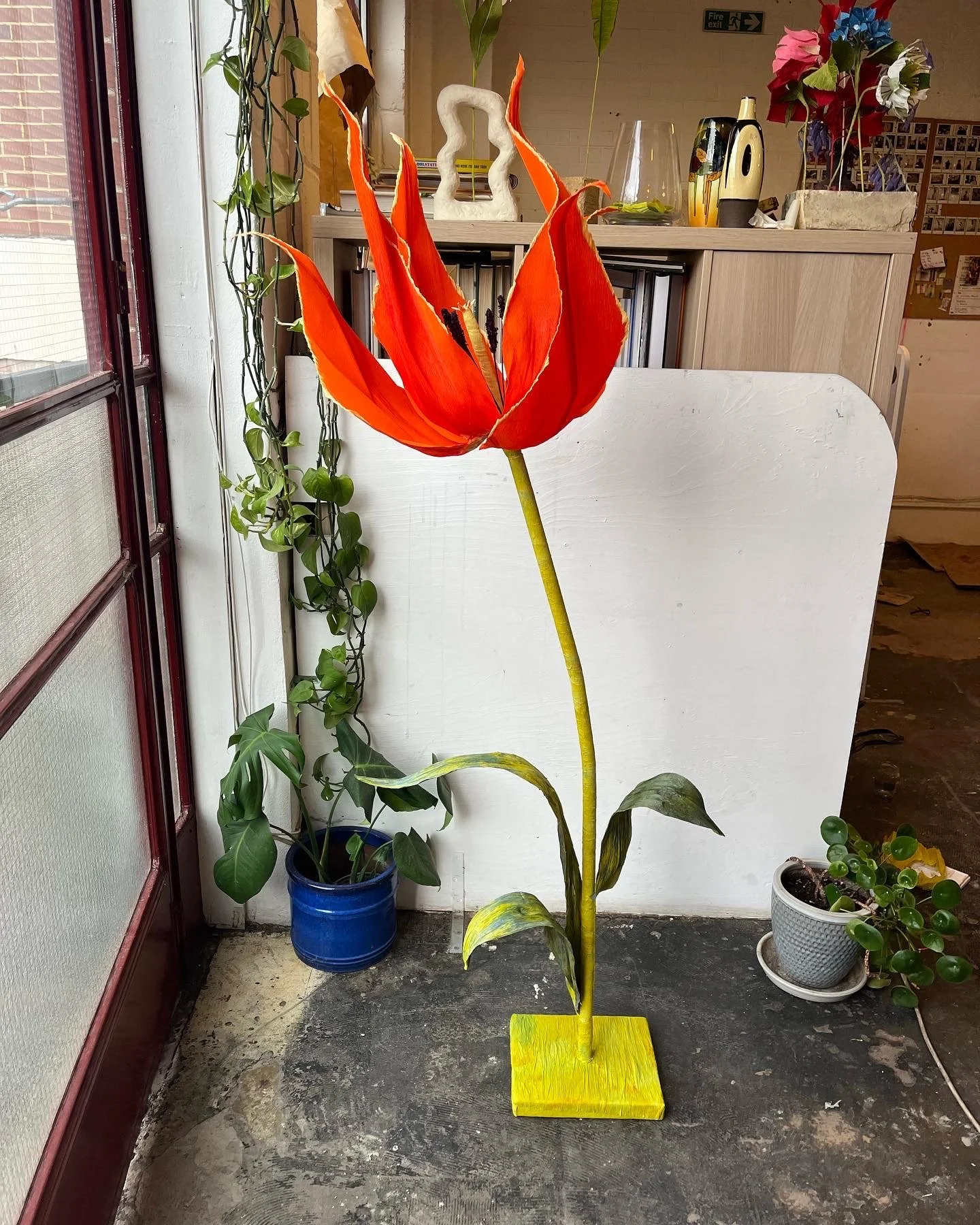 Paper orange and yellow tulip sculpture  by Emmeli Kimhi on  a yellow square base, placed indoors next to potted green plants and near a window.