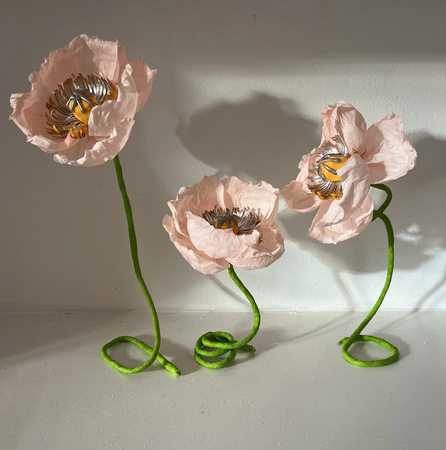 Three artificial pink poppy flowers by Emmeli Kimhi with green wire stems casting shadows on a white wall.