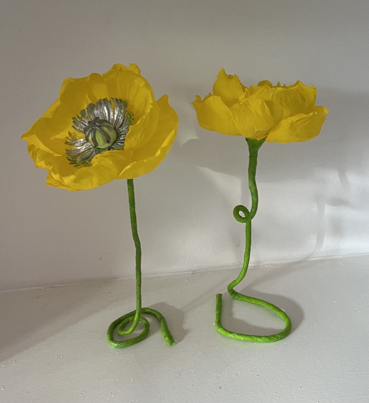 Decorative yellow flowers by Emmeli Kimhi with green wire stems, resembling poppies, on a white background.