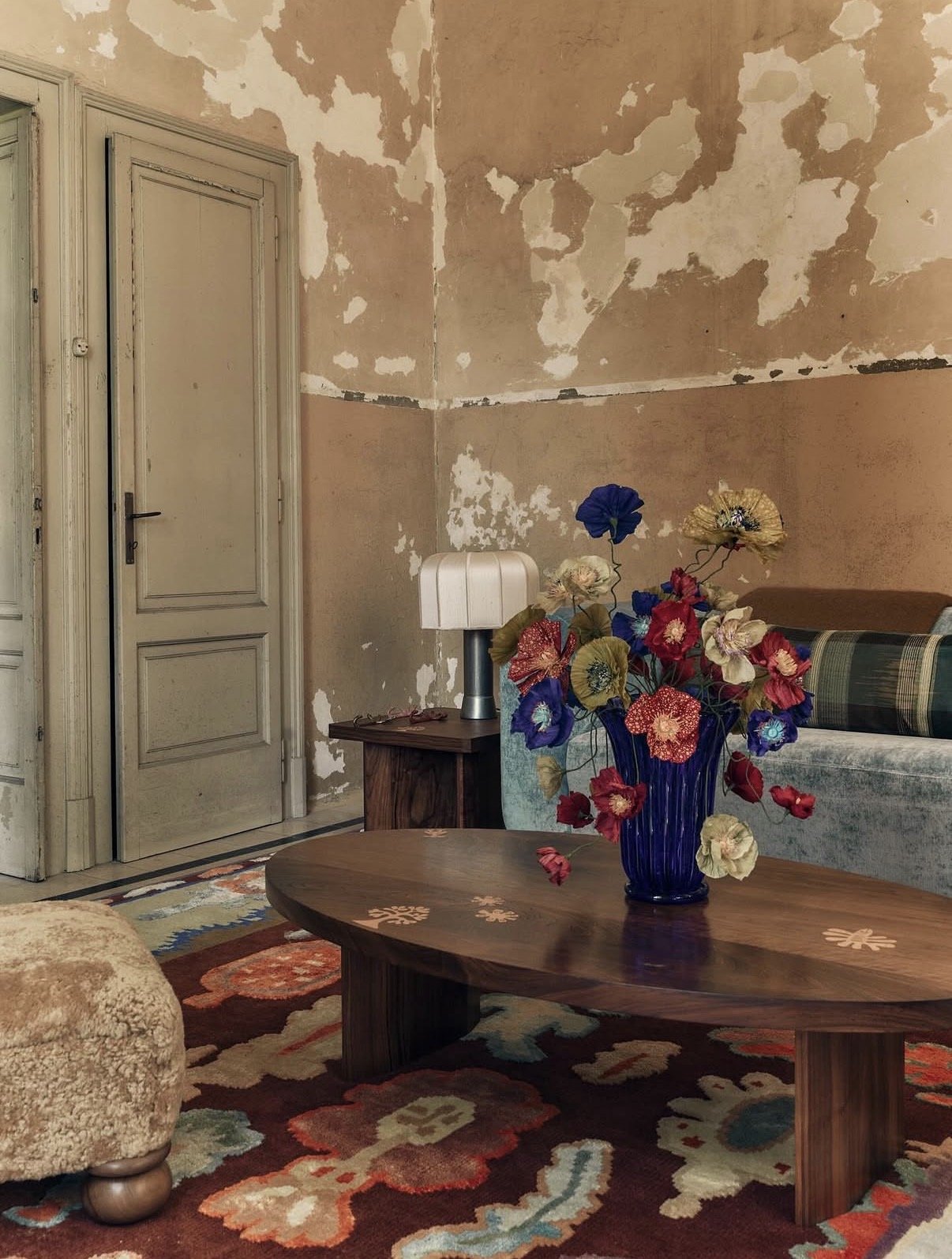 Emmeli Kimhi’s vibrant hand-painted paper poppy bouquet in a blue ribbed glass vase, styled on a wooden oval coffee table. The sculptural arrangement features rich reds, blues, and creams against a backdrop of distressed walls in Studio Ashby’s insta