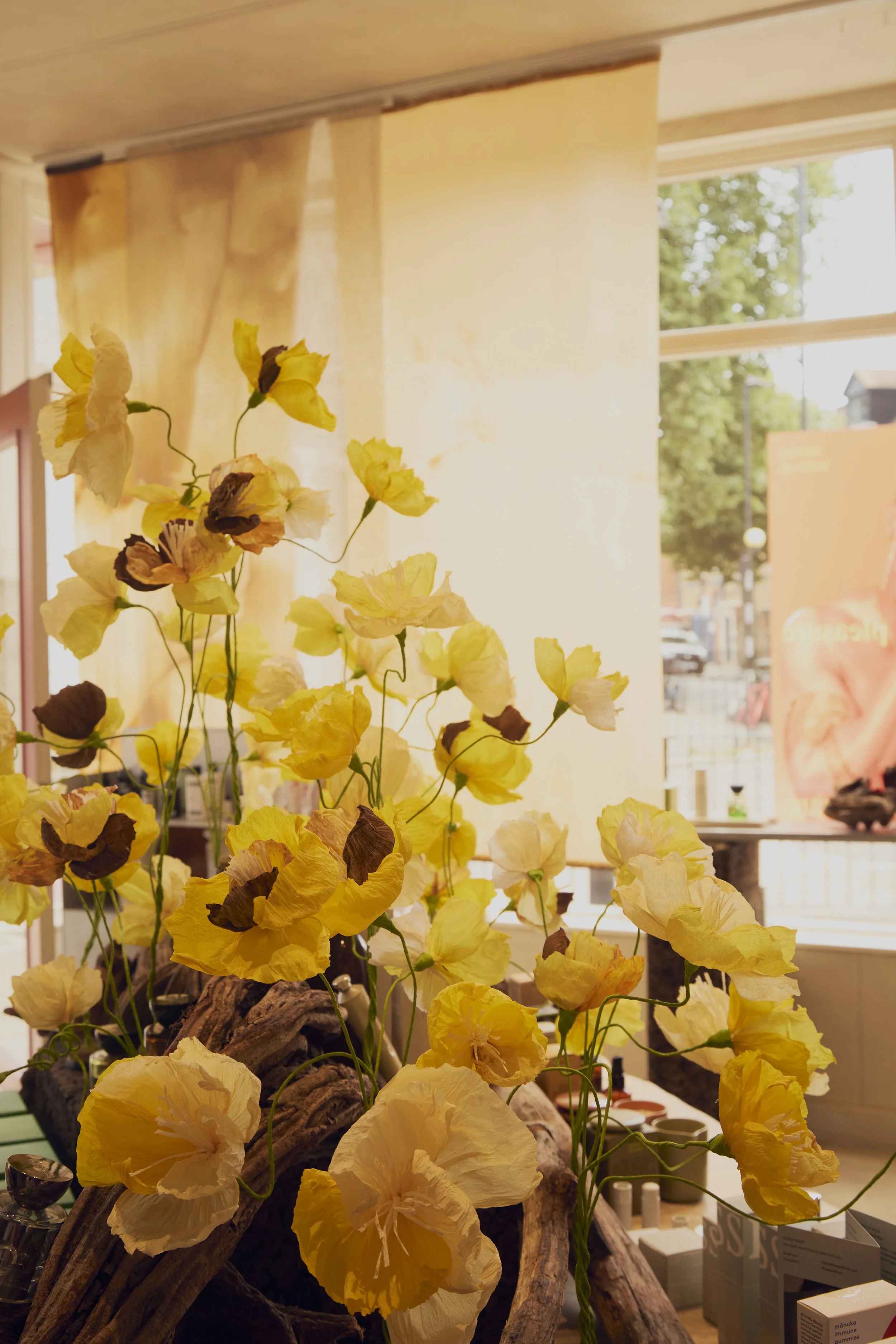 Yellow and brown paper flowers arranged on a decorative piece inside a store with large windows and cream-colored walls.
