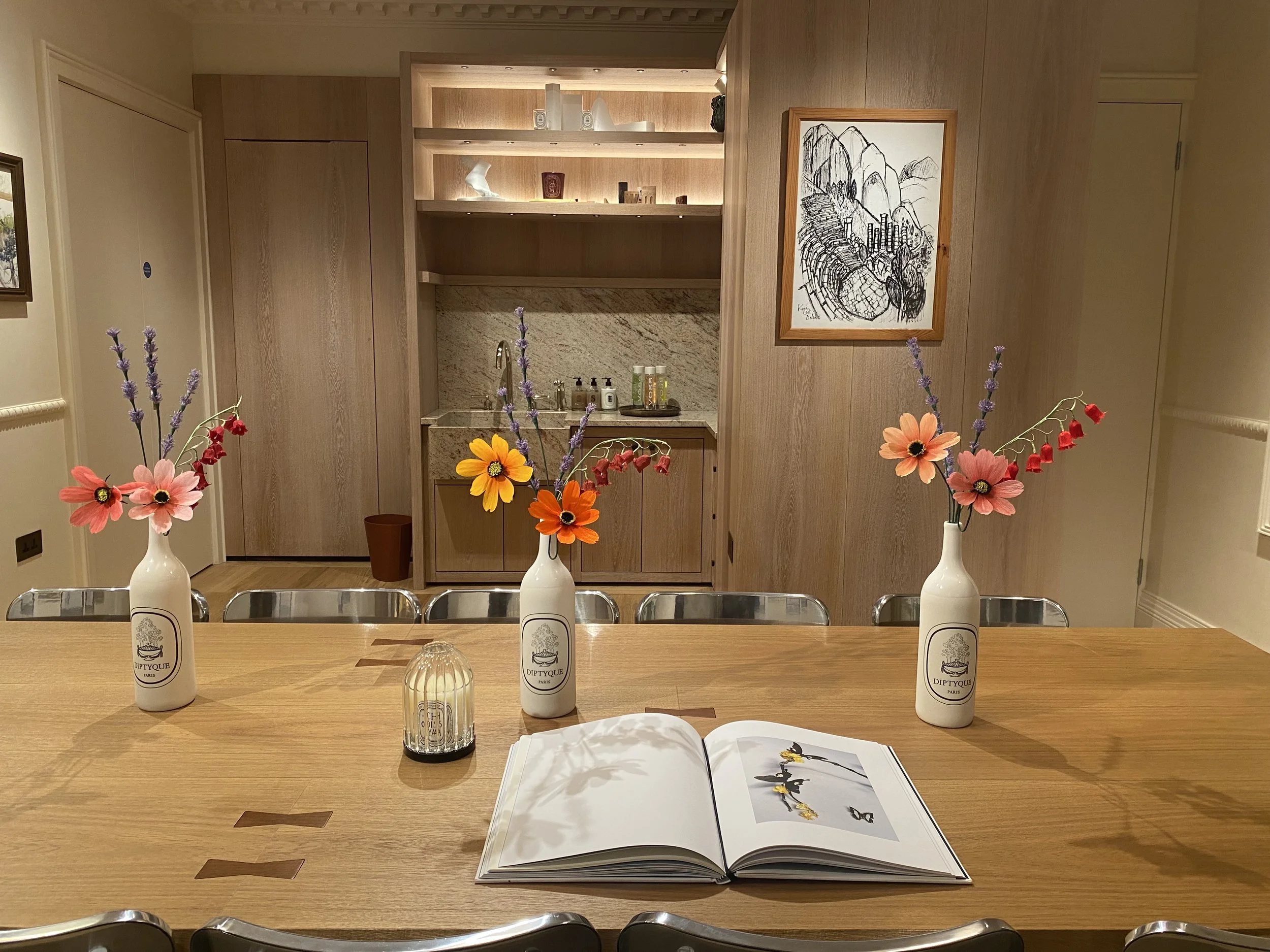 A dining table with four white bottles labeled "Diptyque Paris" and a black and white open book showing a fashion illustration, with three vases of colorful flowers and a clear glass candle holder in the foreground. In the background, a wooden kitchenette with shelving and a framed black-and-white landscape drawing.