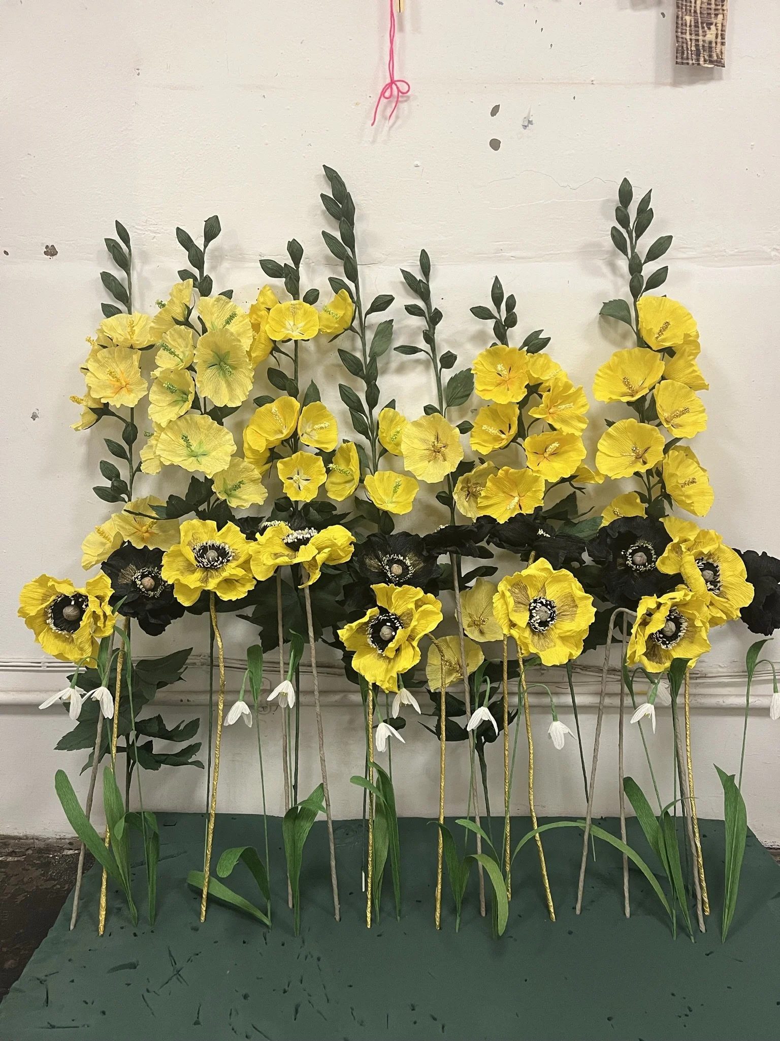 Artificial yellow and black flowers by Emmeli Kimhi arranged on green stems and leaves, displayed against a plain wall.