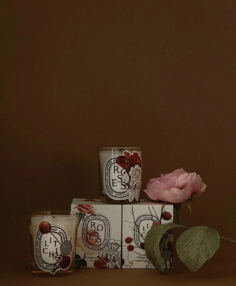 A display of three candles with floral and fruit designs, stacked on boxes, with a pink flower and green leaves by Emmeli Kimhi next to them, against a brown background.
