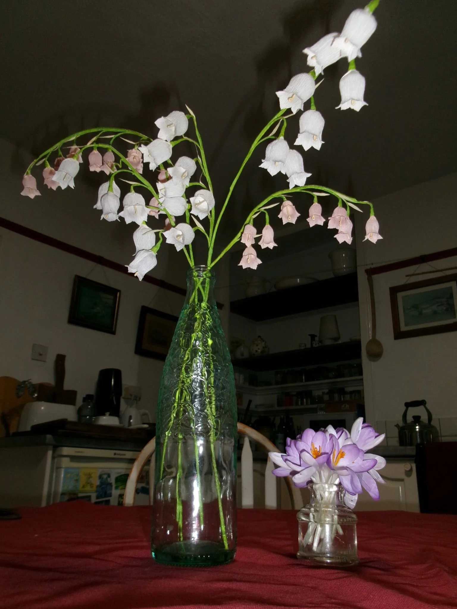 A tall green glass vase with white and pink flowers by Emmeli Kimhi  and elongated green leaves, alongside a small clear glass vase with purple and white crocus flowers, all on a red tablecloth in a room with framed pictures and kitchen shelves.