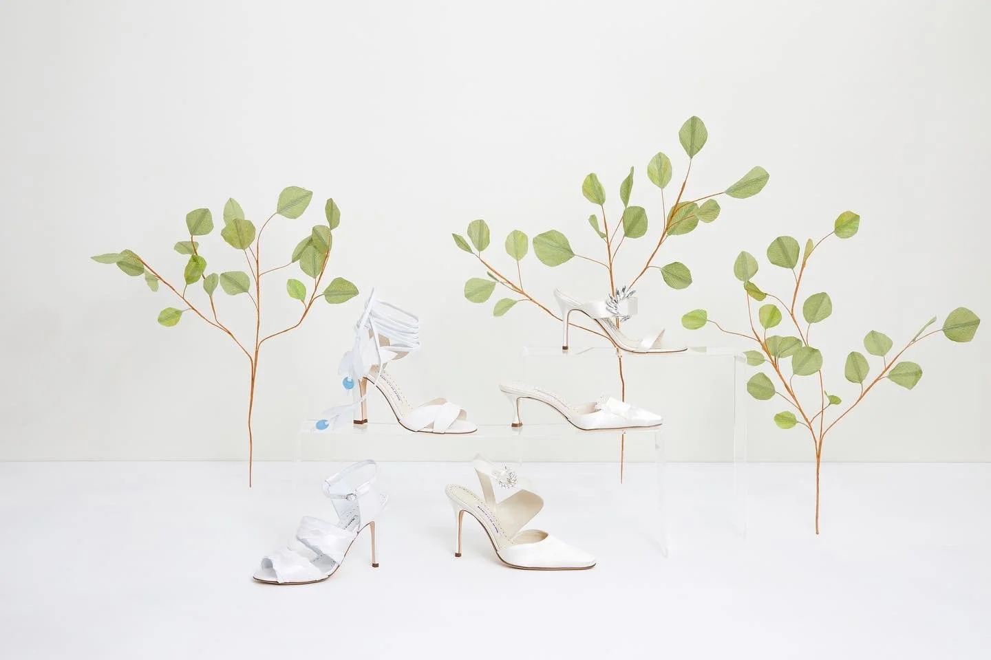 Display of white high-heeled sandals on clear platforms, with decorative paper branches with green leaves by Emmeli Kimhi in the background.
