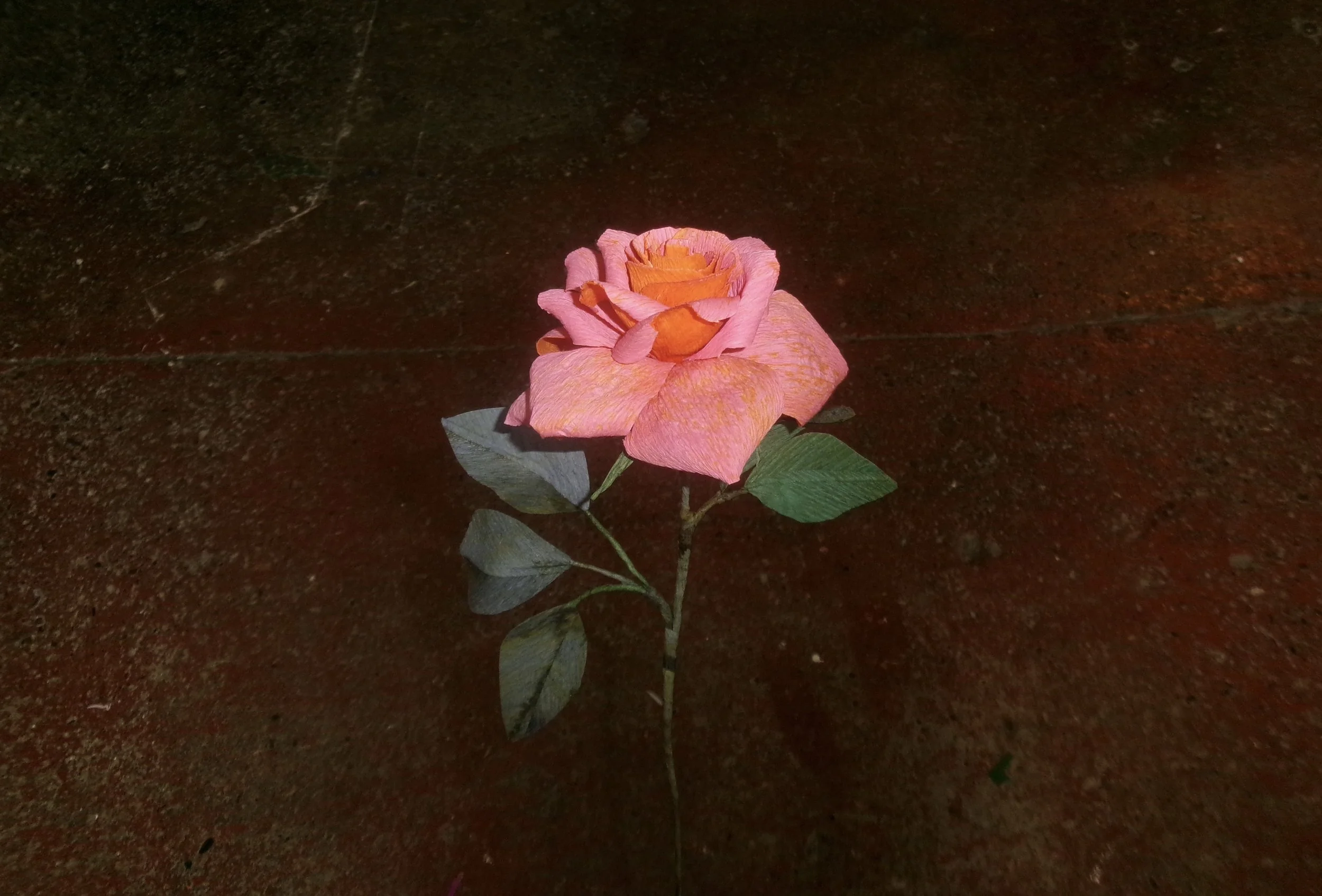 Pink and orange artificial rose by Emmeli Kimhi lying on a dark, textured surface.